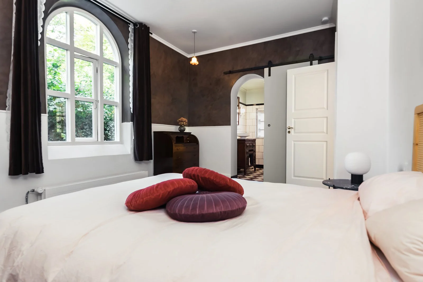 A bedroom with a white bed, four circular red and purple pillows, a black bedside table with a white lamp, a window with black curtains, and a view of a bathroom through an arched doorway.