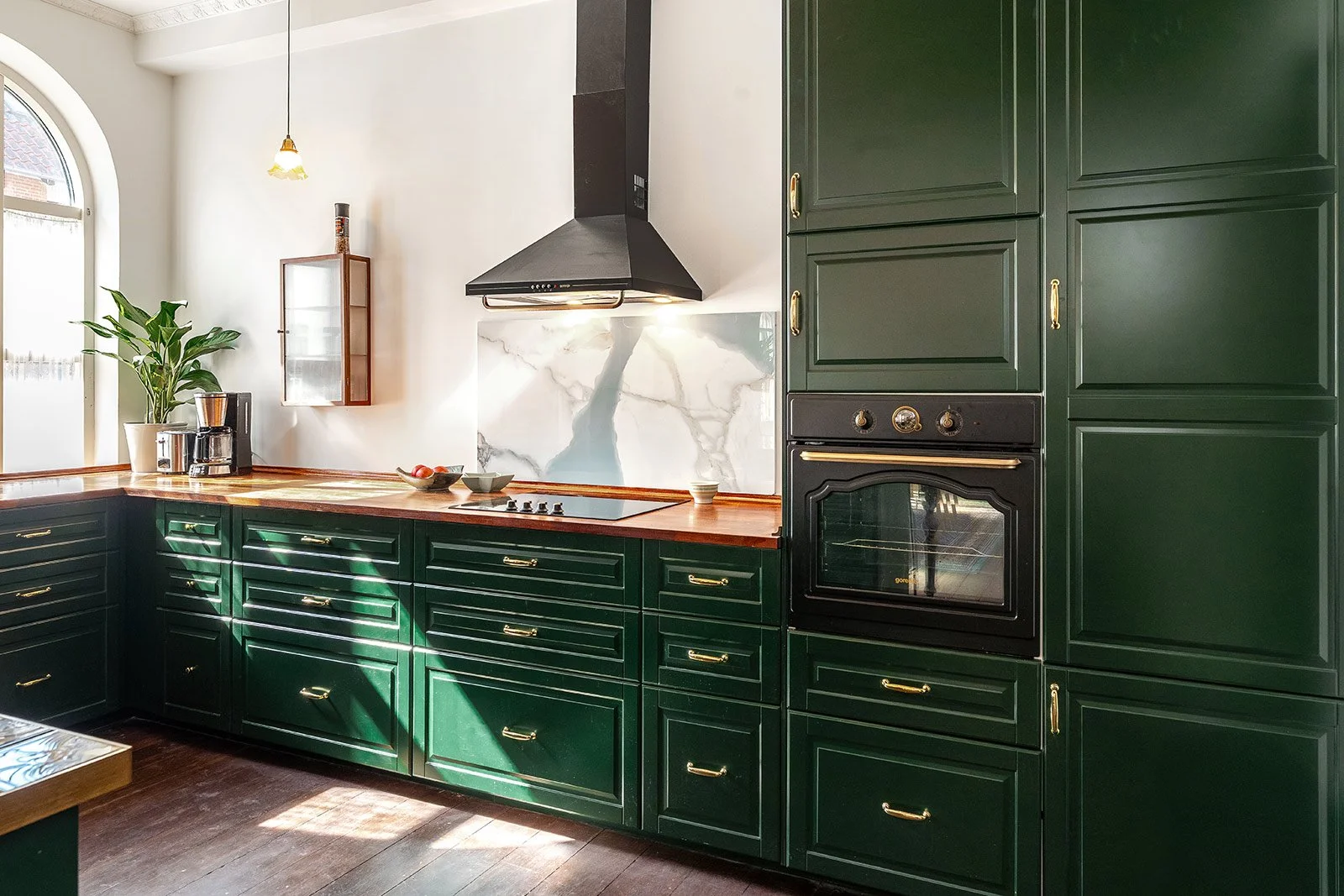 Kitchen with dark green cabinets, wooden countertop, black oven, black range hood, white marble backsplash, large window, potted plant, and hanging light fixture.