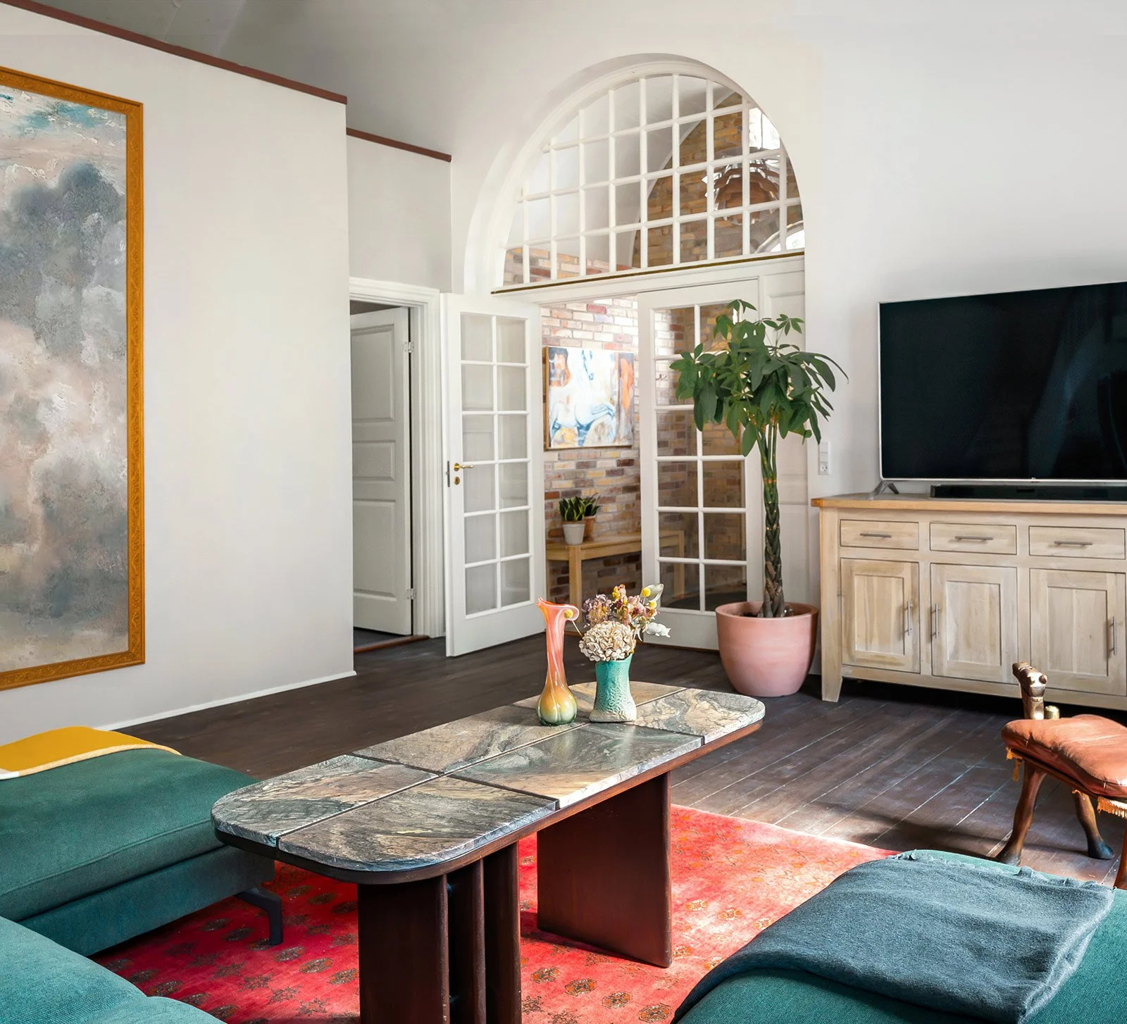 Living room with dark wood flooring, green sofa, coffee table with vases and flowers, large potted plant, flat-screen TV, and decorative artwork, with an arched window and open door leading to a brick-walled space.