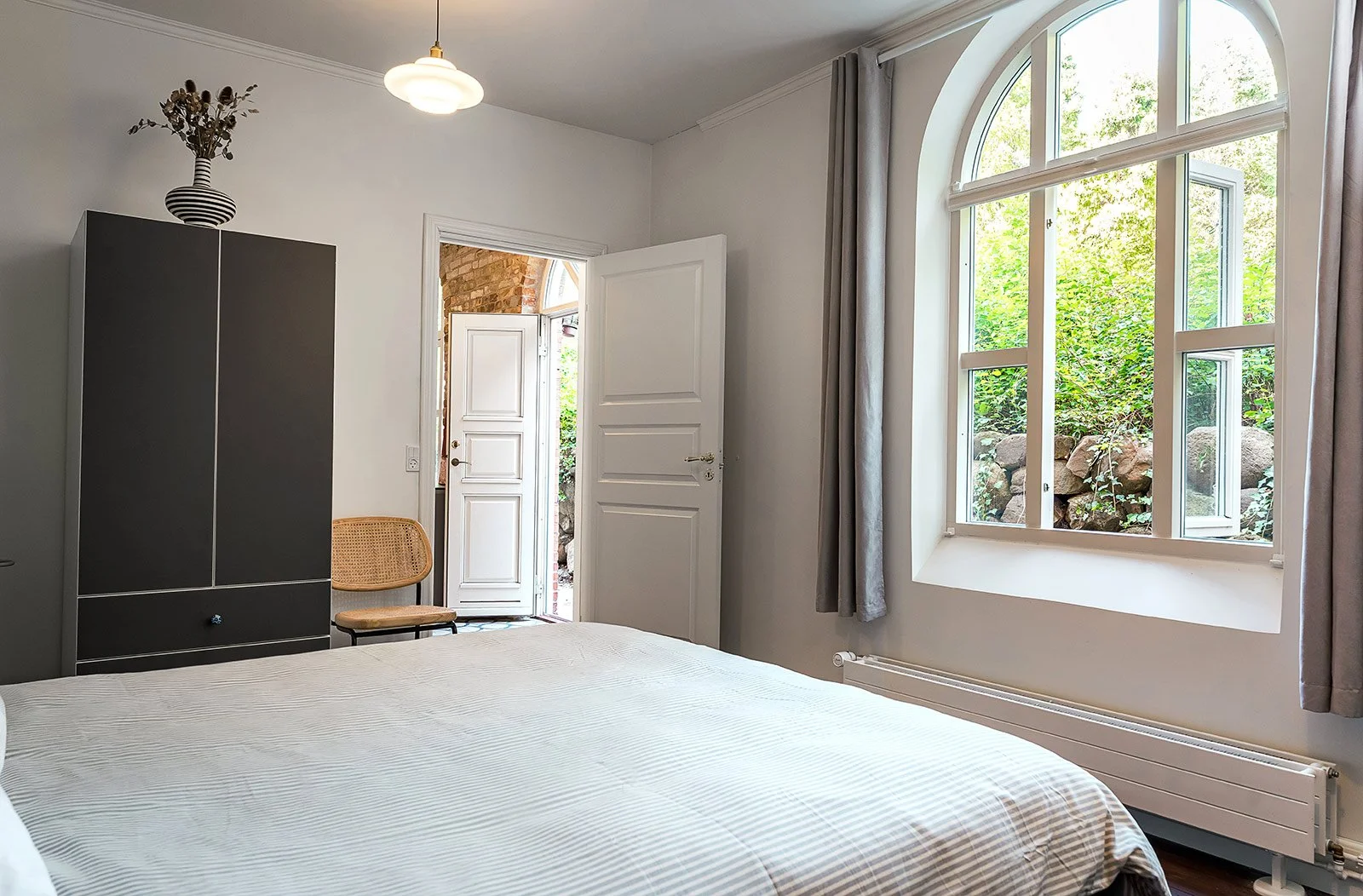 A bedroom with a bed, a large window with light curtains, an open door leading outside, and a black wardrobe topped with a striped vase of dried flowers.
