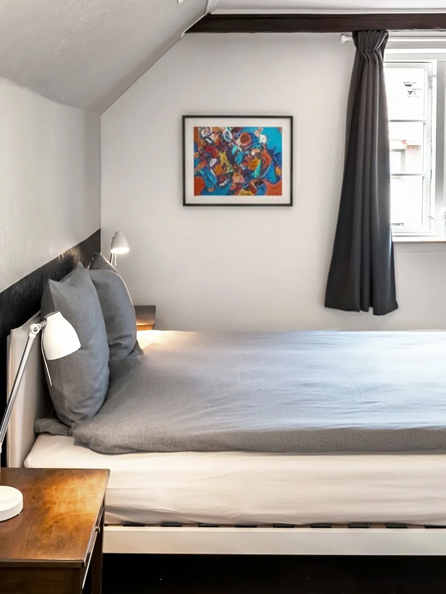 A bedroom with a bed covered in gray sheets and pillows, a wooden nightstand with a white lamp, a window with dark curtains, and a colorful abstract painting on the white wall.