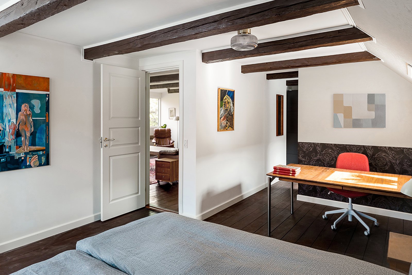 Interior of a bedroom and office space with white walls, wooden beams, contemporary artwork, a bed with light bedding, a desk with red chair, and an open door leading to a living room with armchair and window.