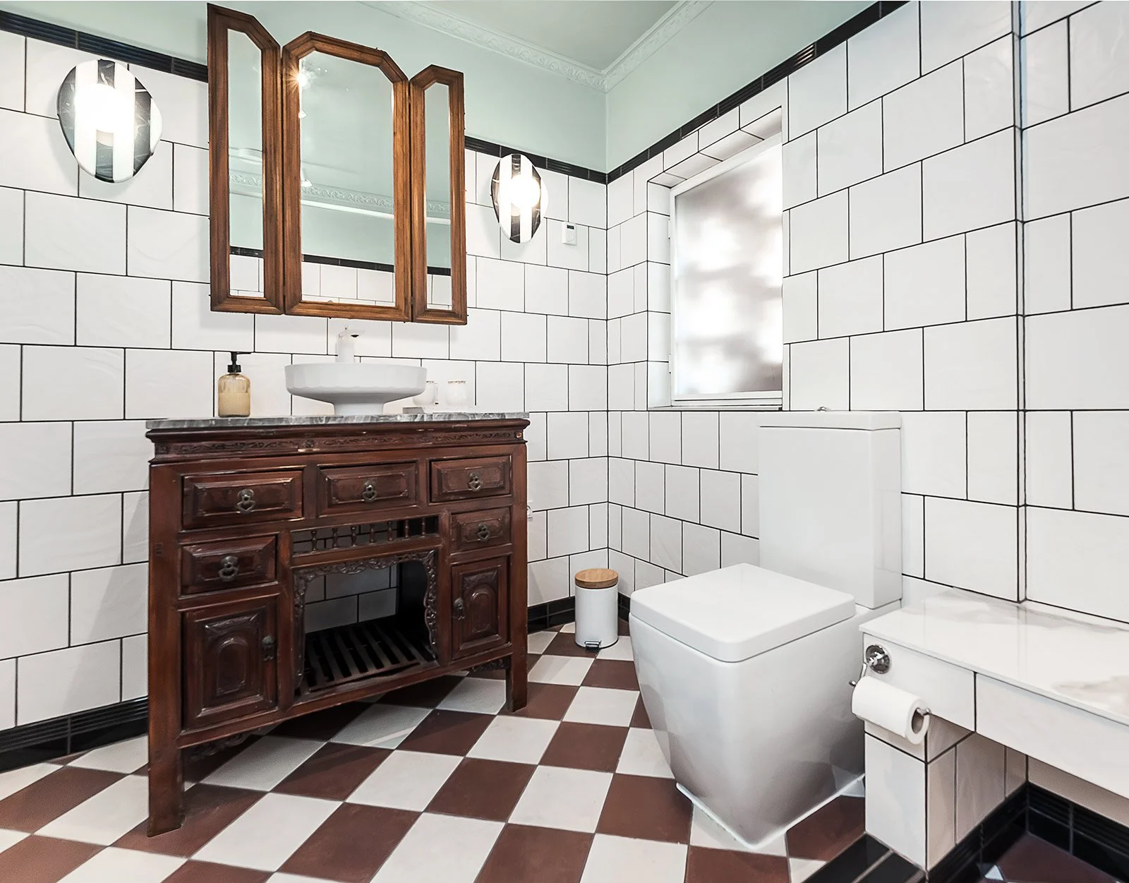 Bathroom with a white toilet, a wooden vanity with a vessel sink, a mirror cabinet, and wall-mounted lights, with white tiled walls and a checkered brown and white floor.