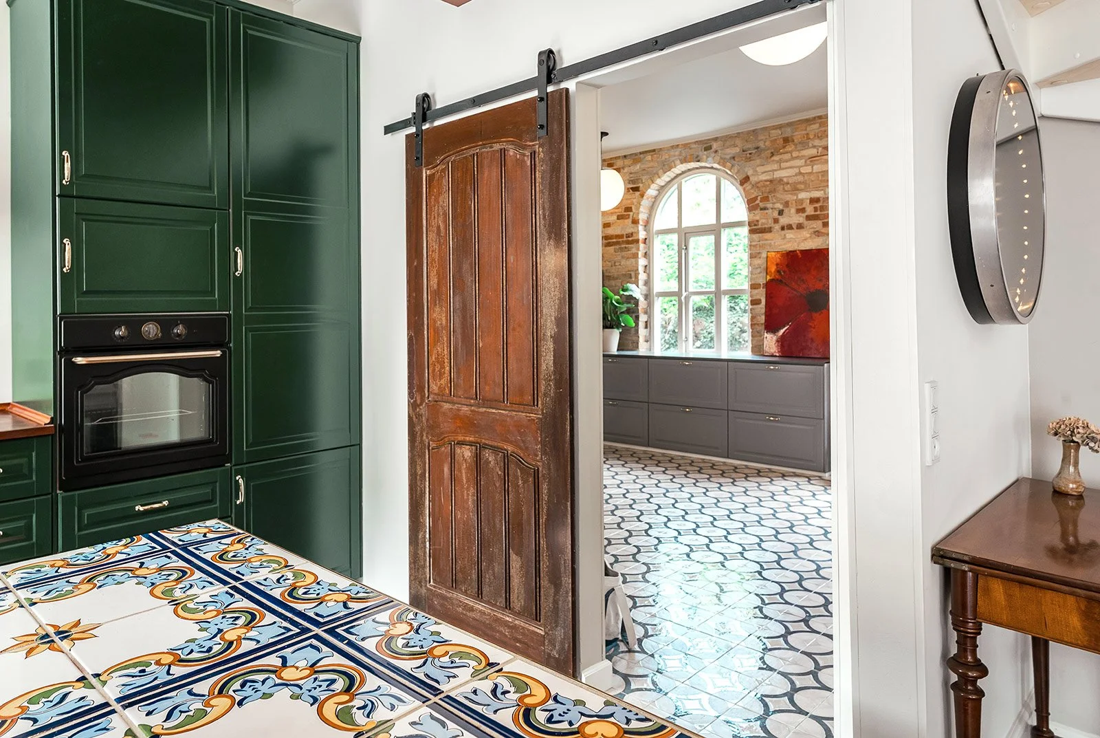 View of a kitchen with dark green cabinets, a patterned tiled table, a sliding wooden barn door, and an adjacent room with brick walls, a large arched window, gray cabinets, potted plant, and abstract painting.