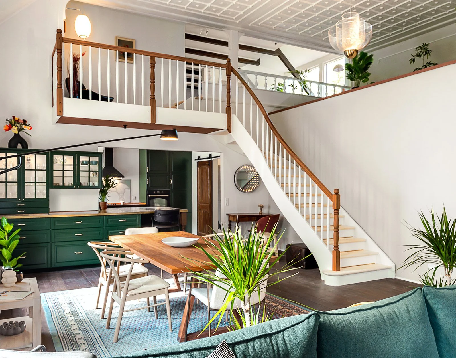 Interior of a modern townhouse with an open-plan living and dining area, green kitchen cabinets, a dining table with white chairs, and a staircase with wooden handrails leading to an upstairs balcony.