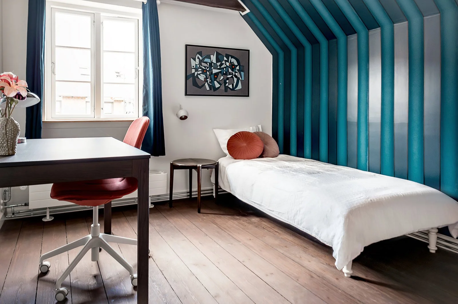 A bedroom with a single bed with white bedding and decorative pillows, a wooden chair next to a small side table, a window with blue curtains, a desk with a red chair, and a blue textured wall backdrop behind the bed. There's modern artwork on the wa