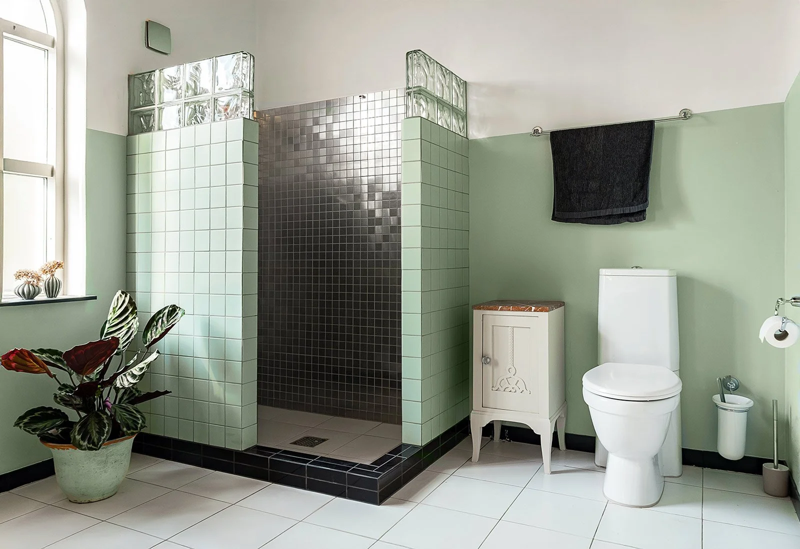 A modern bathroom with a small shower area with black tiles, a white toilet, a small white cabinet, a black towel hanging on a rod, a large window with sunlight, a potted plant, and a white tile floor.