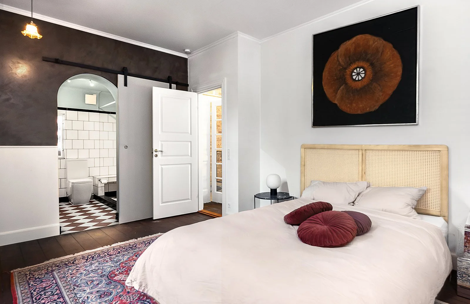 A bedroom with a bed that has a woven rattan headboard, white bedding, and decorative pillows. A black nightstand with a round white lamp is beside the bed. A large, framed artwork of an orange-brown spiral shell hangs above the bed. An open door rev