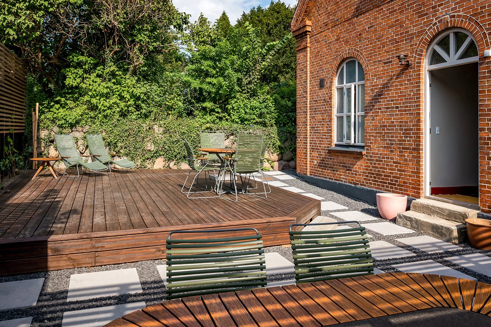 A backyard patio area with a wooden deck, outdoor chairs, and tables, surrounded by greenery and a red brick building with arched windows.