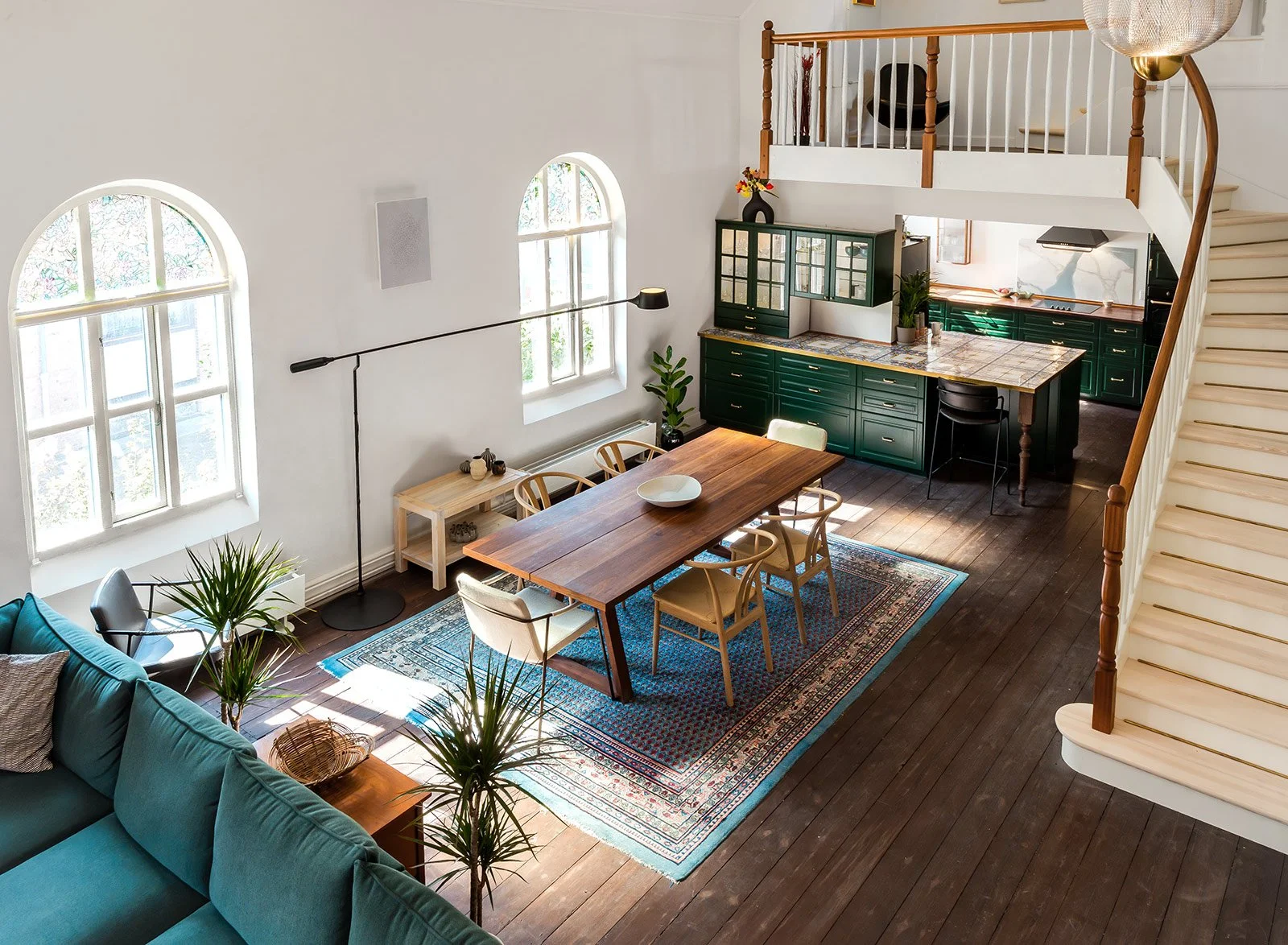 An open-concept living area with a dining space, kitchen, and stairs. Large arched windows let in natural light. There is a wooden dining table with chairs, a green kitchen with cabinets, and a staircase with a wooden handrail.