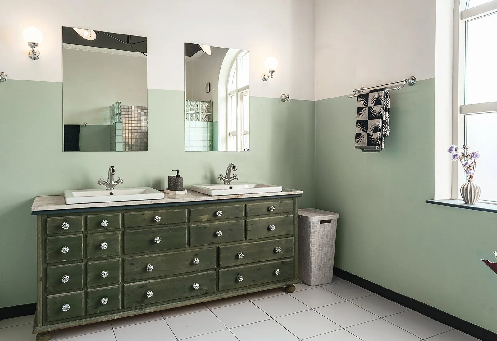 Bathroom with double sink vanity, green cabinet, two mirrors, and window with vase of flowers.