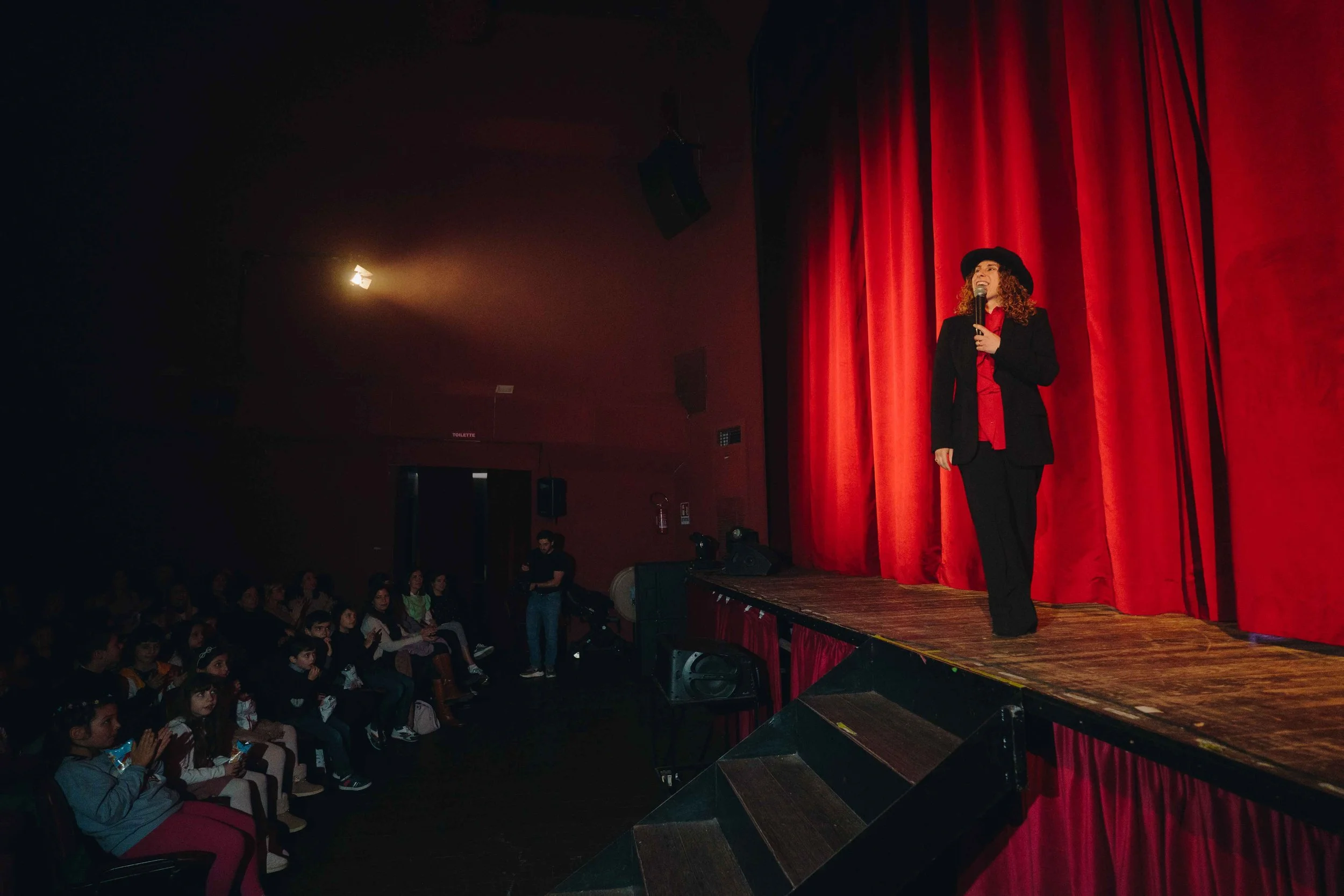 A woman on stage wearing a black hat and suit, speaking into a microphone in front of red curtains, with an audience seated in front of the stage.