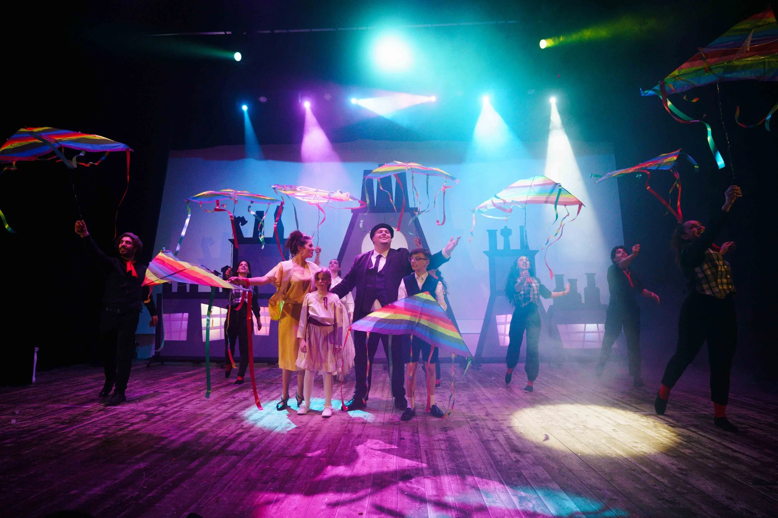 Theater stage with actors holding colorful umbrellas, performing with vibrant lighting and set design of rooftops and chimneys.