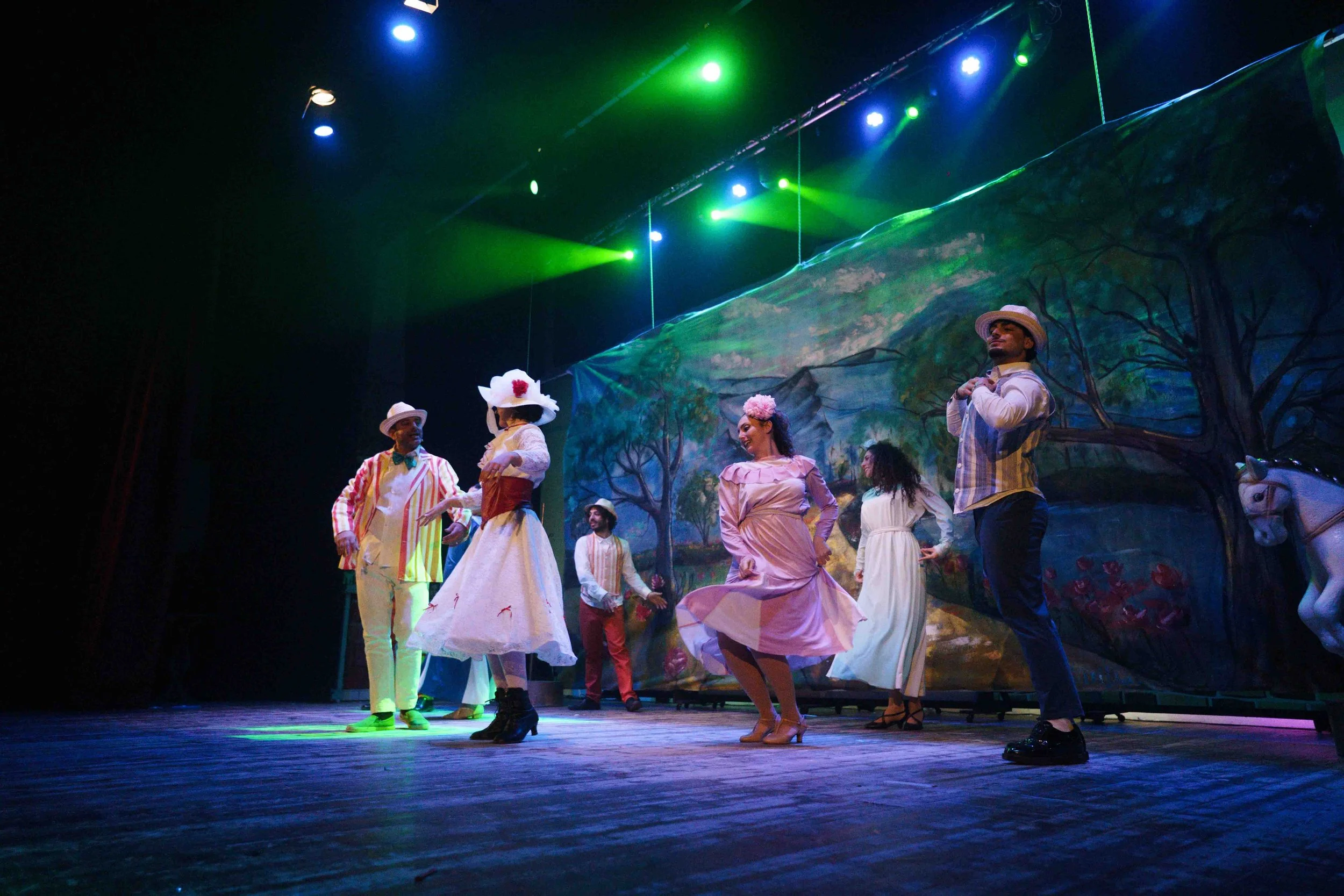 Theatrical performance on stage featuring several actors in vintage costumes, with actors dancing or interacting, set against a scenic background depicting trees and outdoor scenery, illuminated by colorful stage lights.