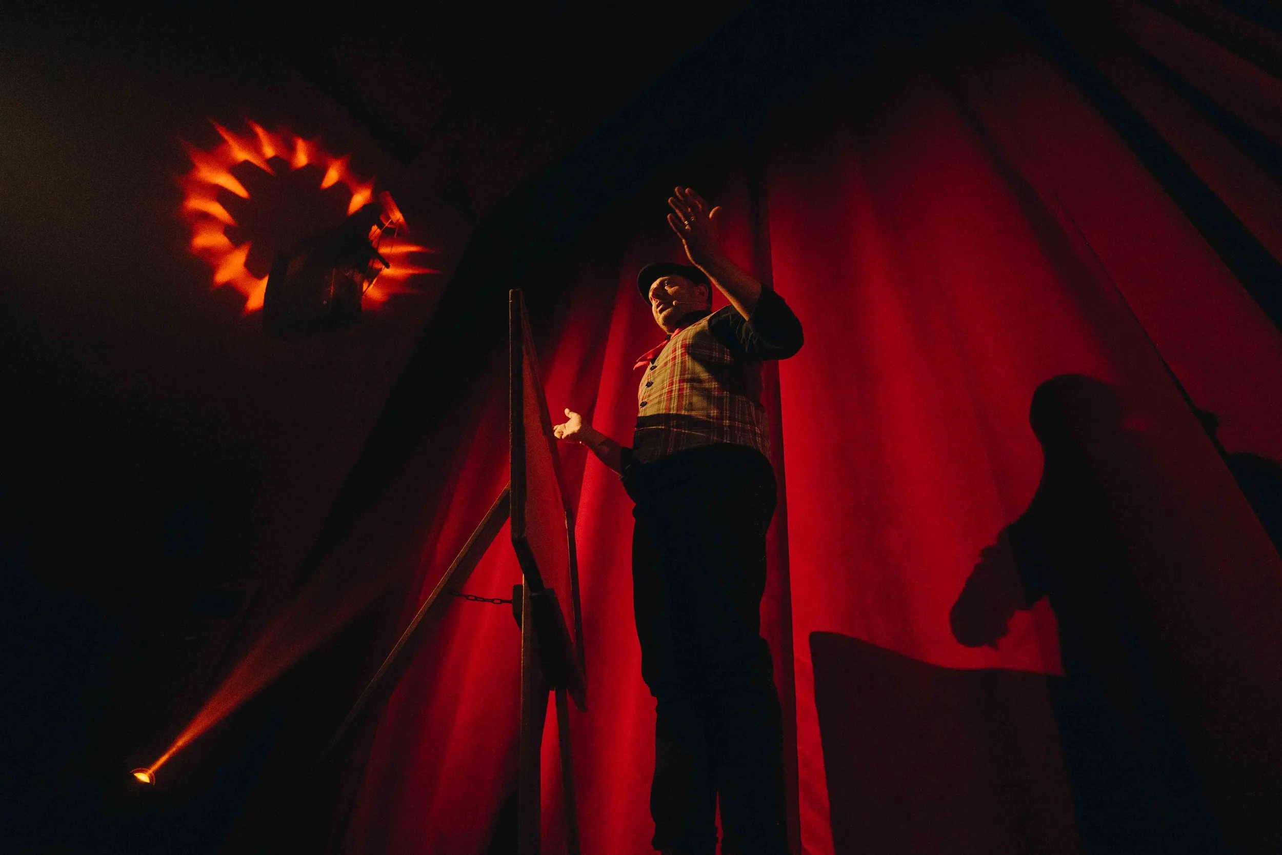 A performer, possibly a magician, stands on stage with a red curtain in the background. The performer is wearing a checkered vest and a black hat, with their right hand raised. There is a spotlight shining towards the ceiling creating a shadow patter