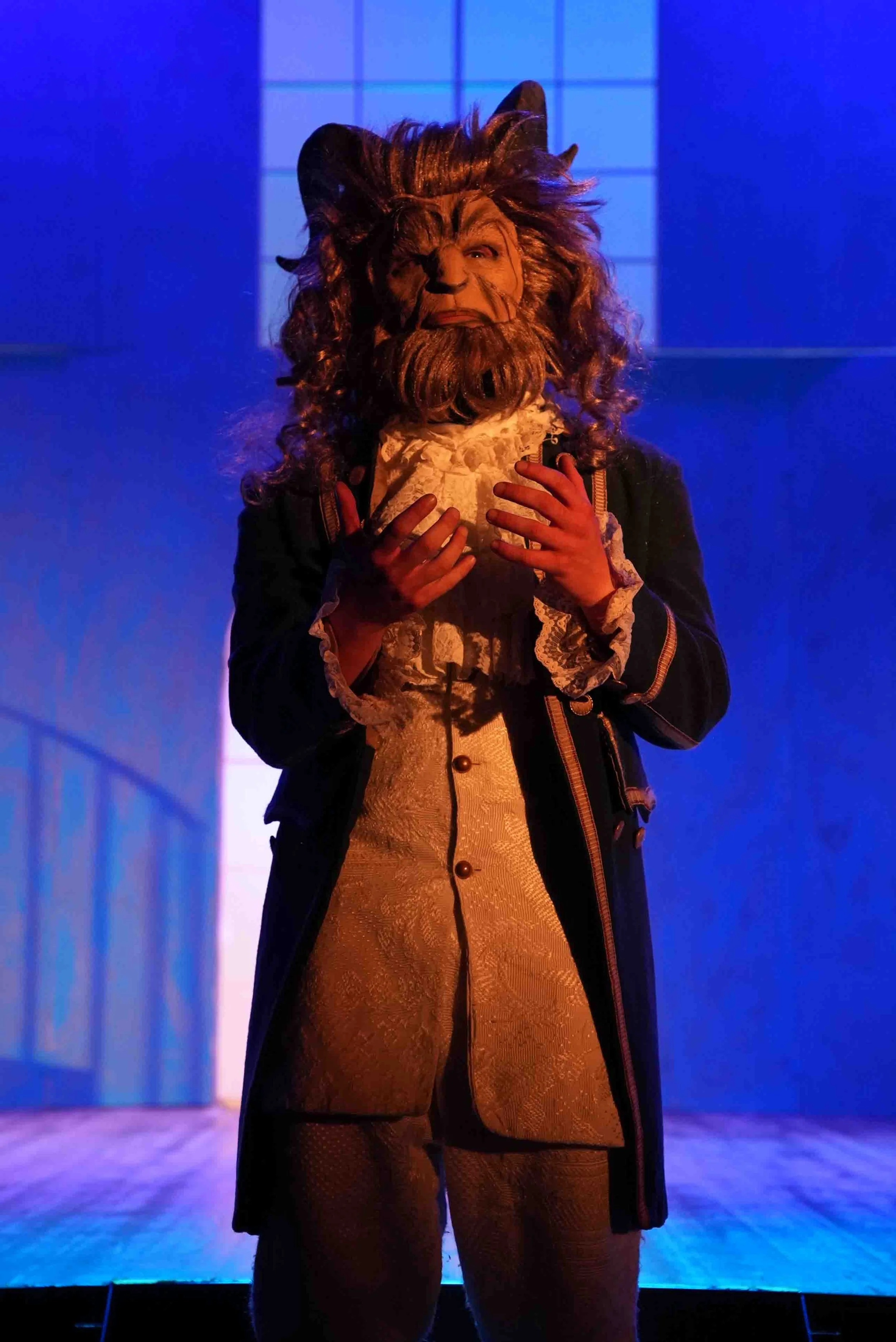 A person in a Beast costume from Beauty and the Beast, wearing a lion mask with a luxurious outfit, standing on a stage with blue lighting.