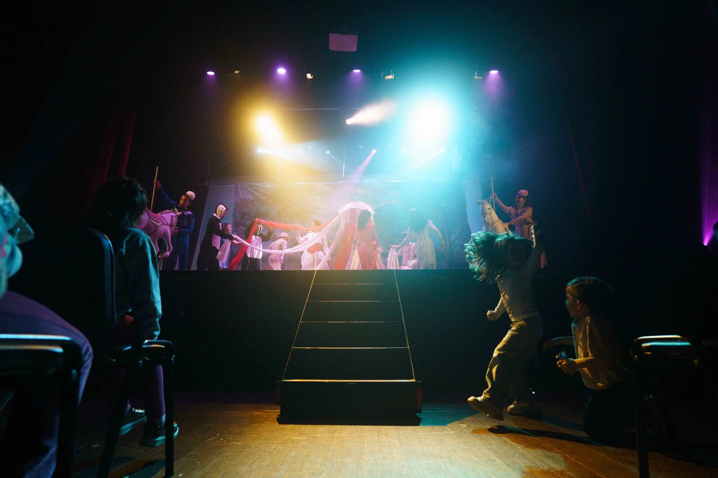 Children watching a colorful stage performance of a circus or theatrical show, with performers in costumes and props, under stage lighting.