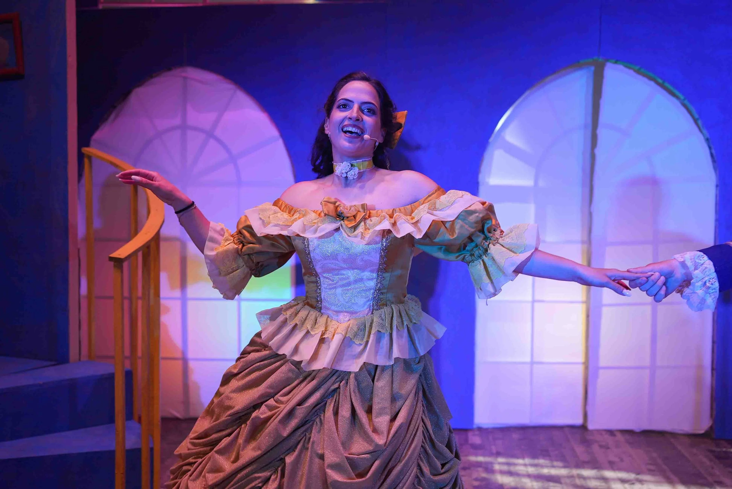 A woman in an elaborate, historical costume singing and holding hands during a stage performance, with a colorful background including large windows and a staircase.