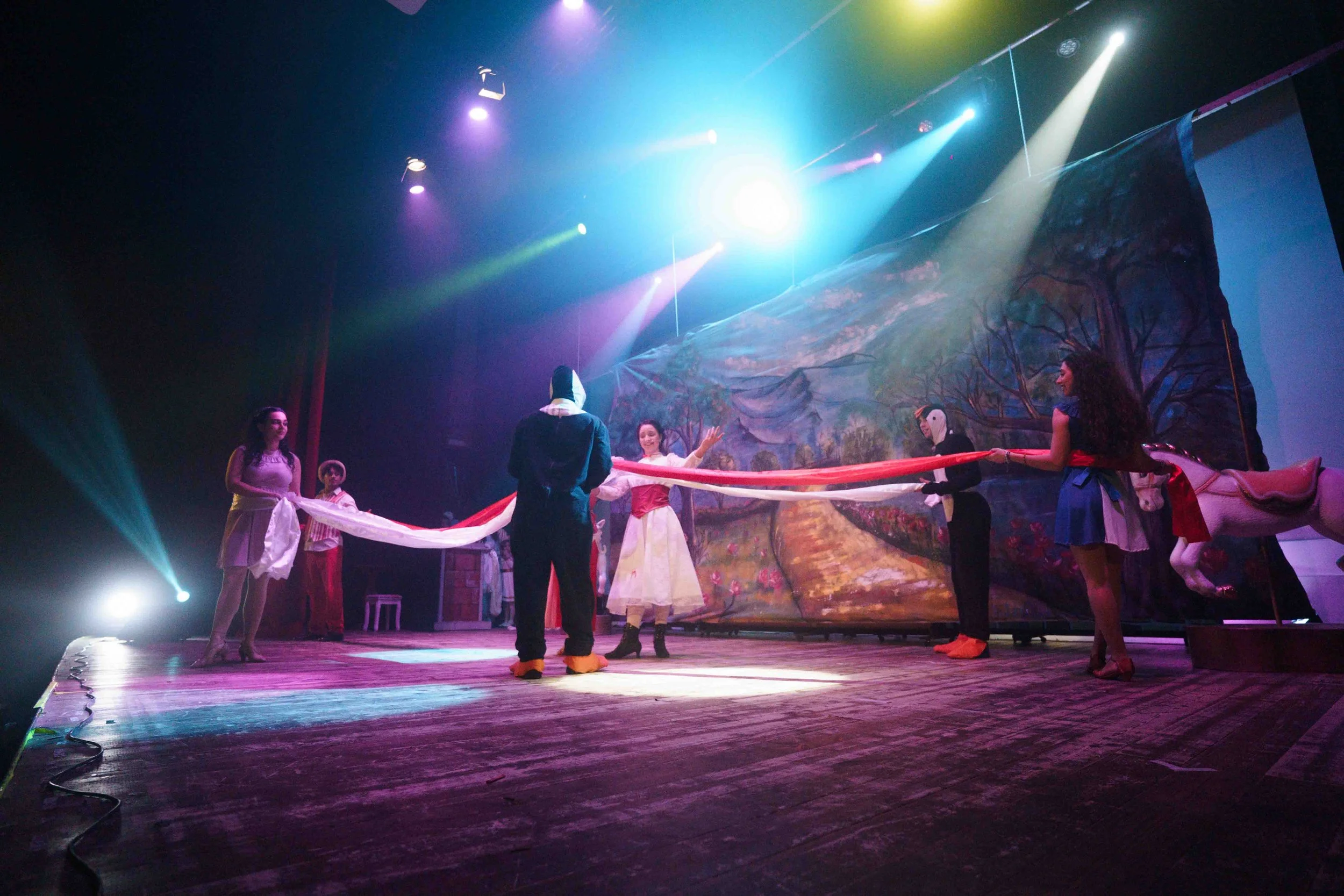 Theatre performers on stage rehearsing a scene with a backdrop painting, stage lighting, and a large prop horse. The actors are holding a long cloth or ribbon.