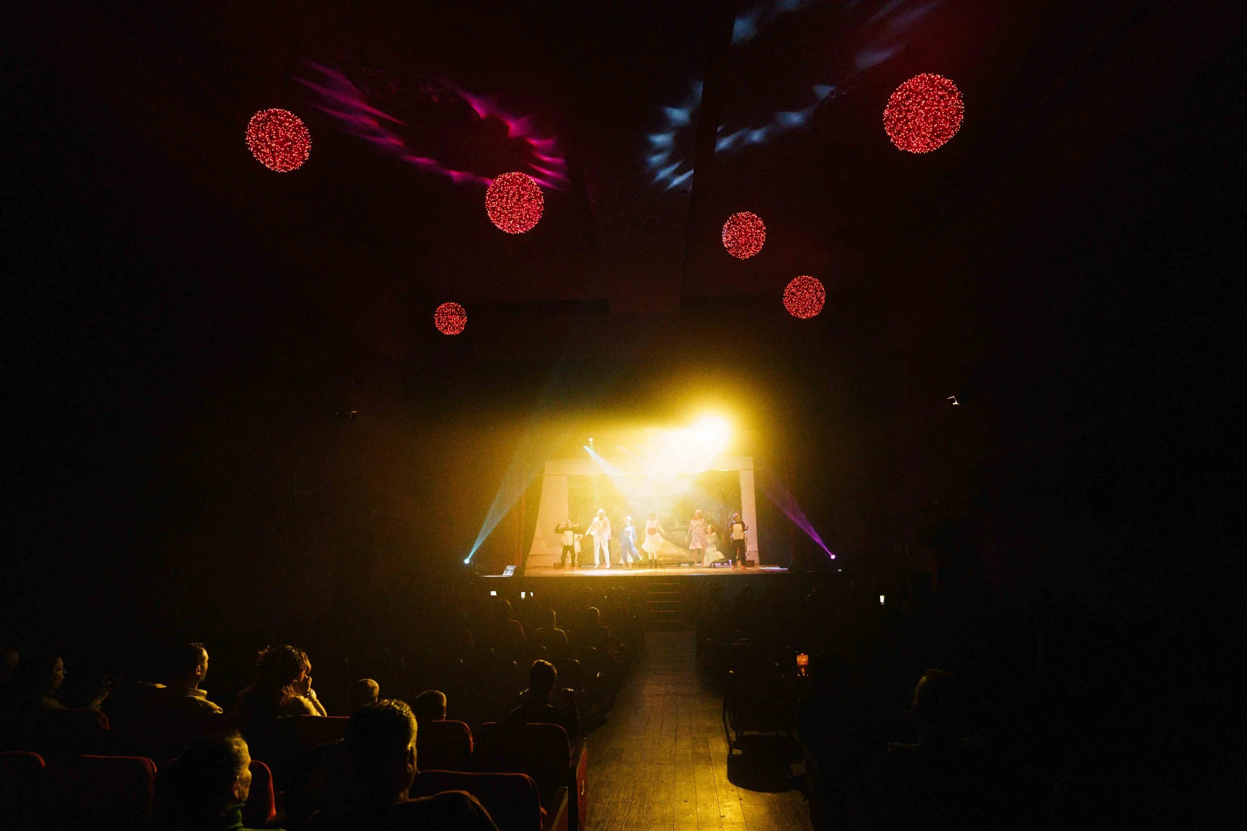 The image shows a stage performance with actors in costumes, illuminated by yellow, purple, and blue stage lights. The audience is seated in a dark theater, with red and purple decorative lights hanging from the ceiling.