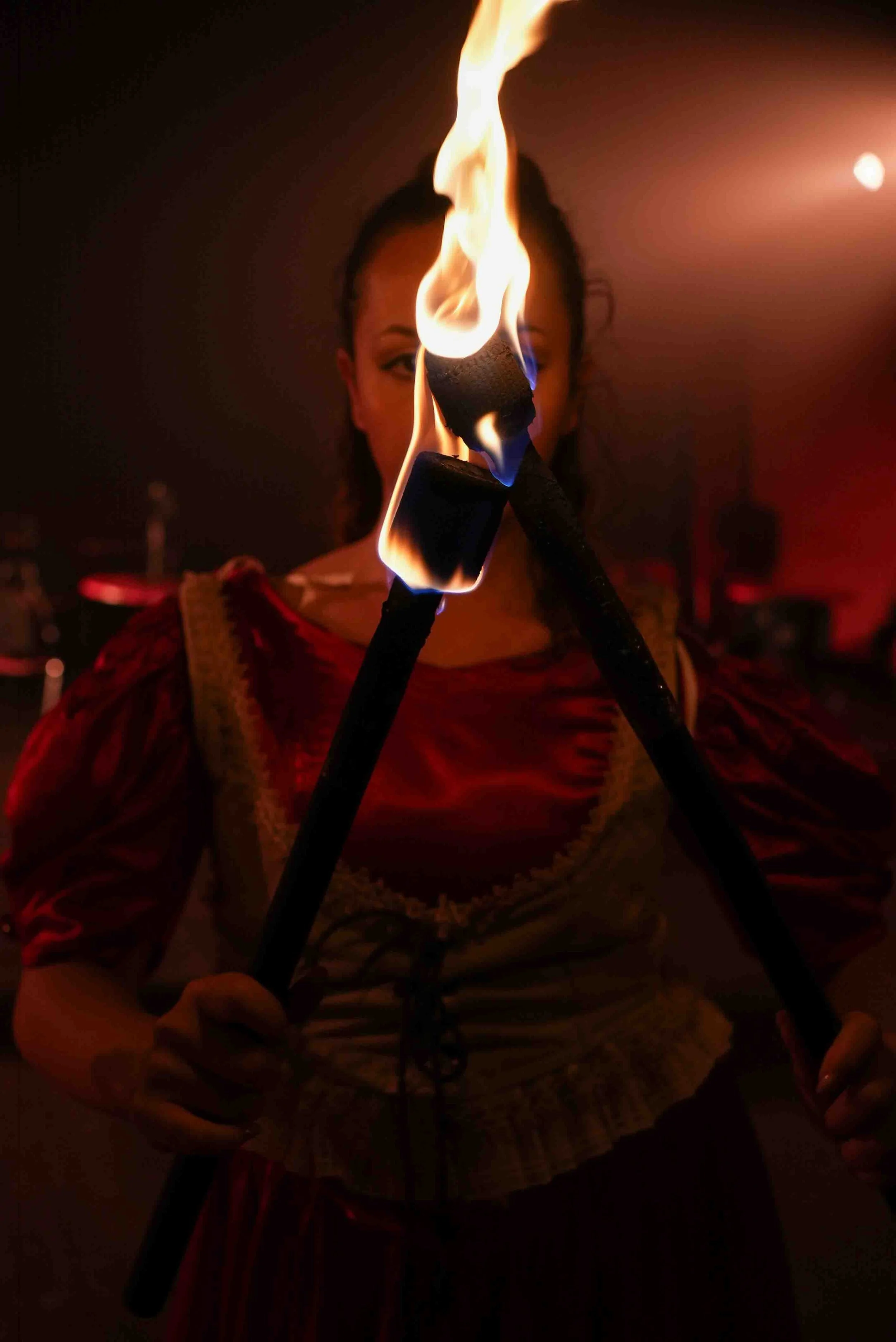 A woman holding two flaming torches in front of her, with the flames obscuring part of her face, in a dark setting.