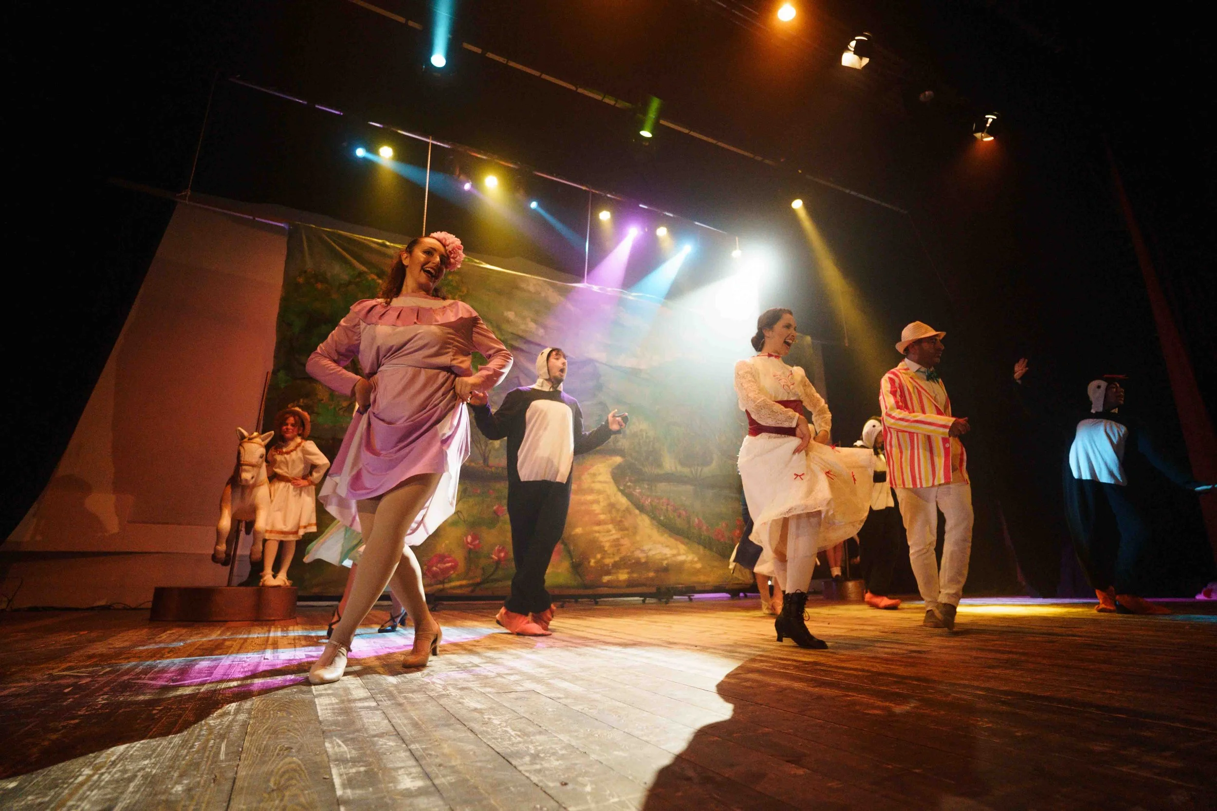 Theater performers on stage in colorful costumes, dancing and singing with a painted scenic backdrop, stage lighting overhead, and wooden stage floor.