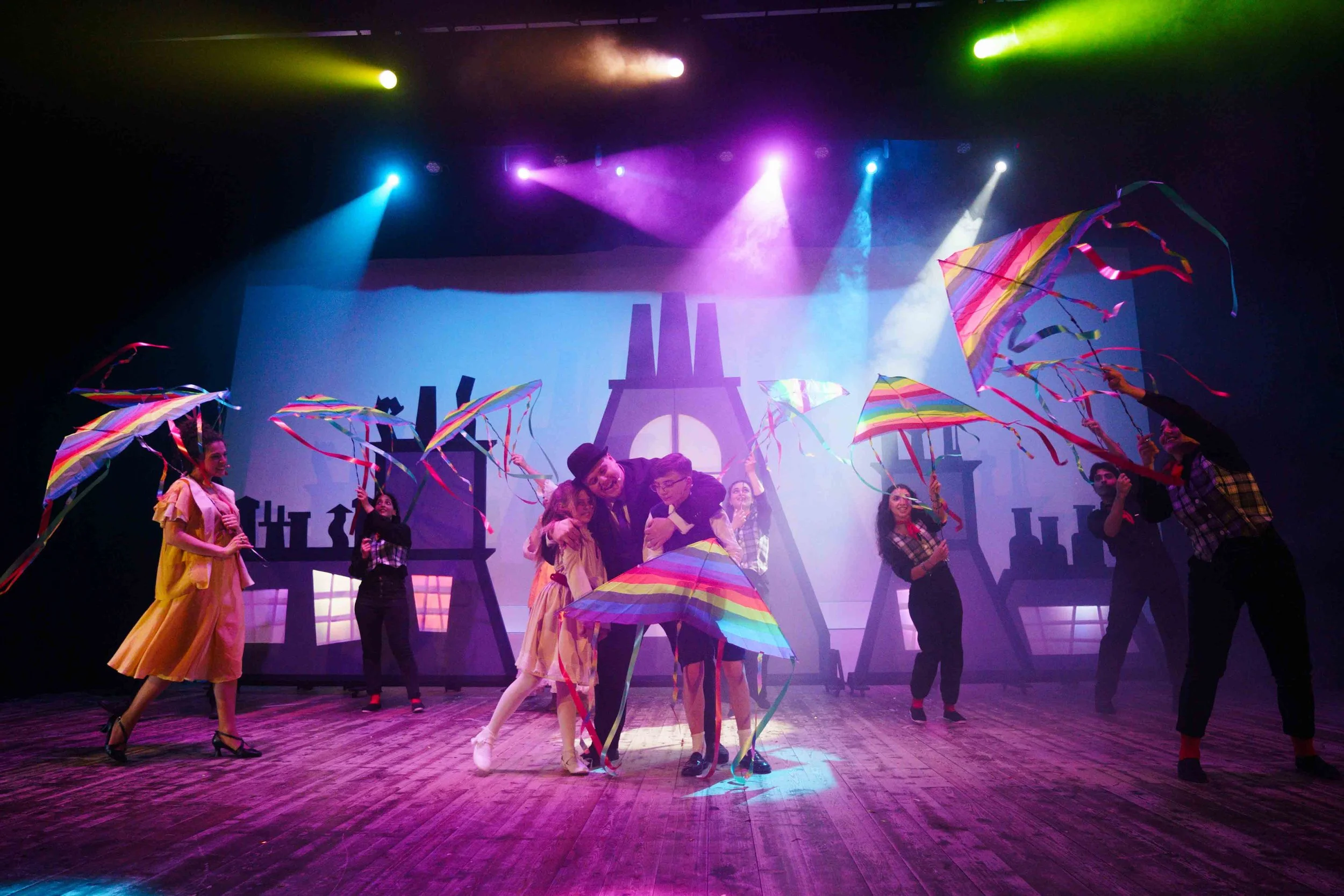 A theatrical stage performance with actors holding colorful umbrellas, under vibrant lighting, with a backdrop resembling city buildings.