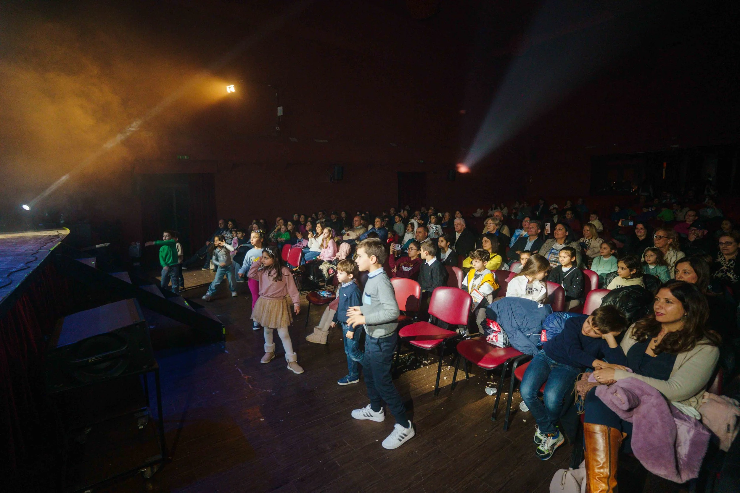 Audience of children and adults sitting in theater seats watching a stage with children performing in front, stage lights shining.