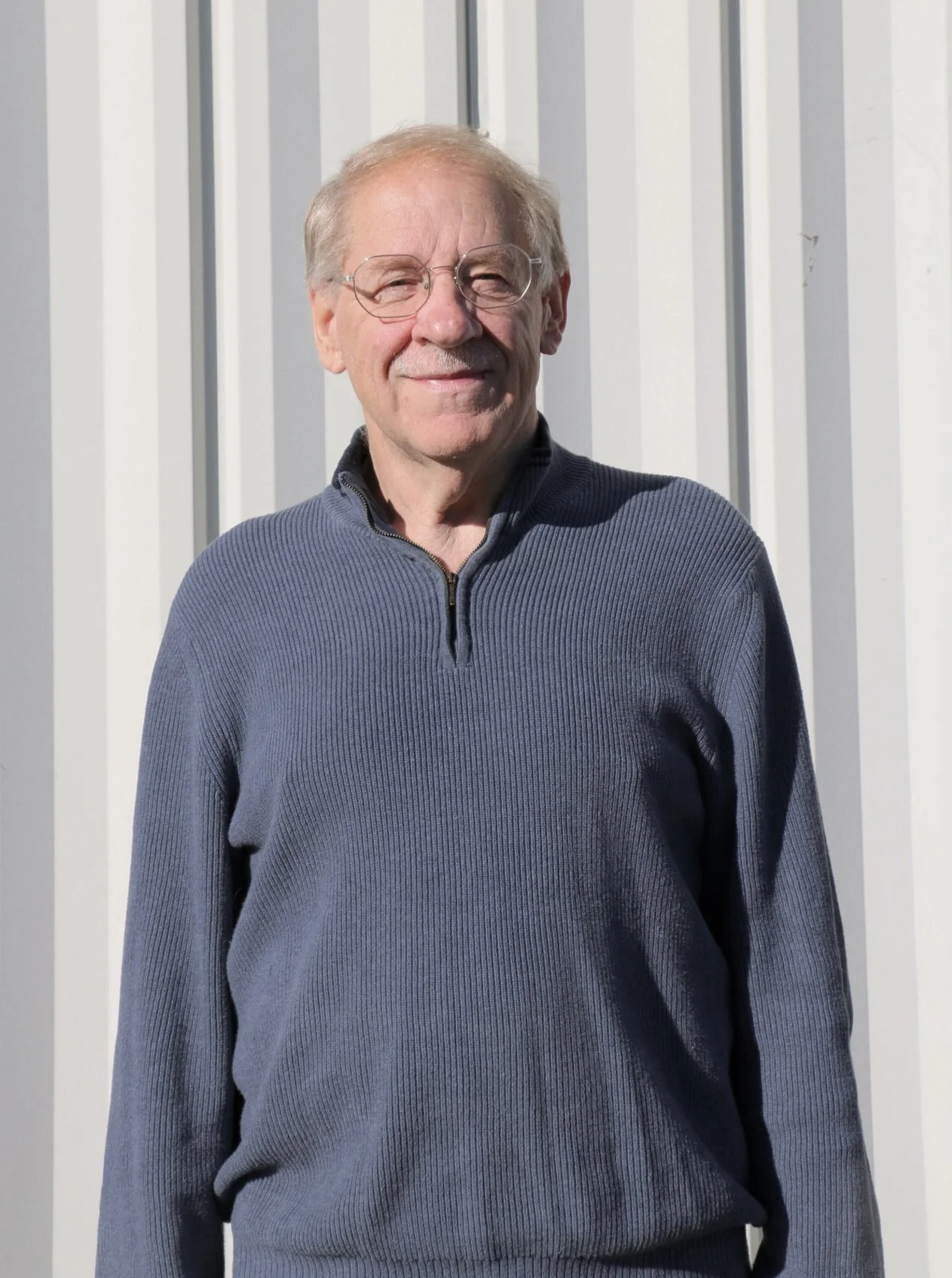 An older man with gray hair, glasses, and a mustache, wearing a gray plaid blazer over a blue checkered shirt, standing against a plain beige wall.