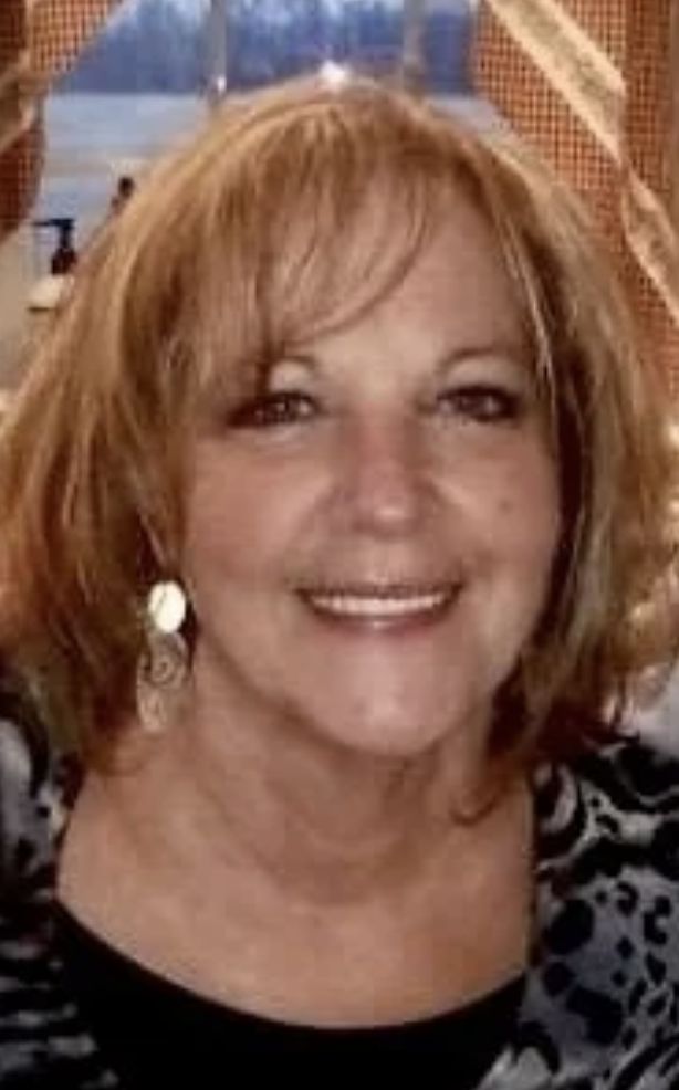 A woman with shoulder-length reddish-brown hair, smiling, wearing earrings and a black and white animal print top, indoors with a window and curtains in the background.