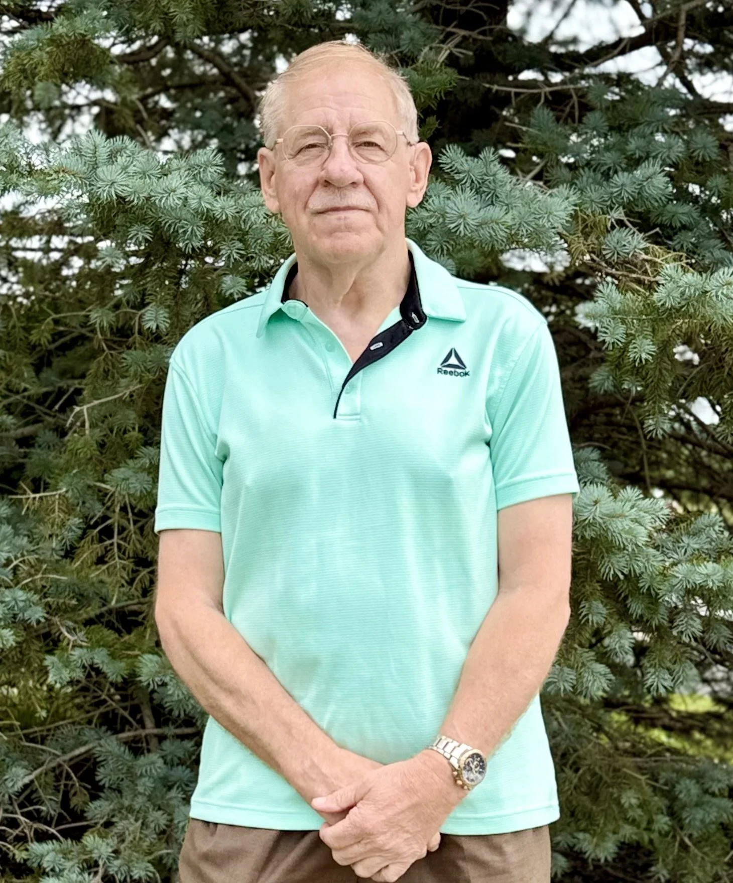 An older man with gray hair, glasses, and a mustache, wearing a gray plaid blazer over a blue checkered shirt, standing against a plain beige wall.