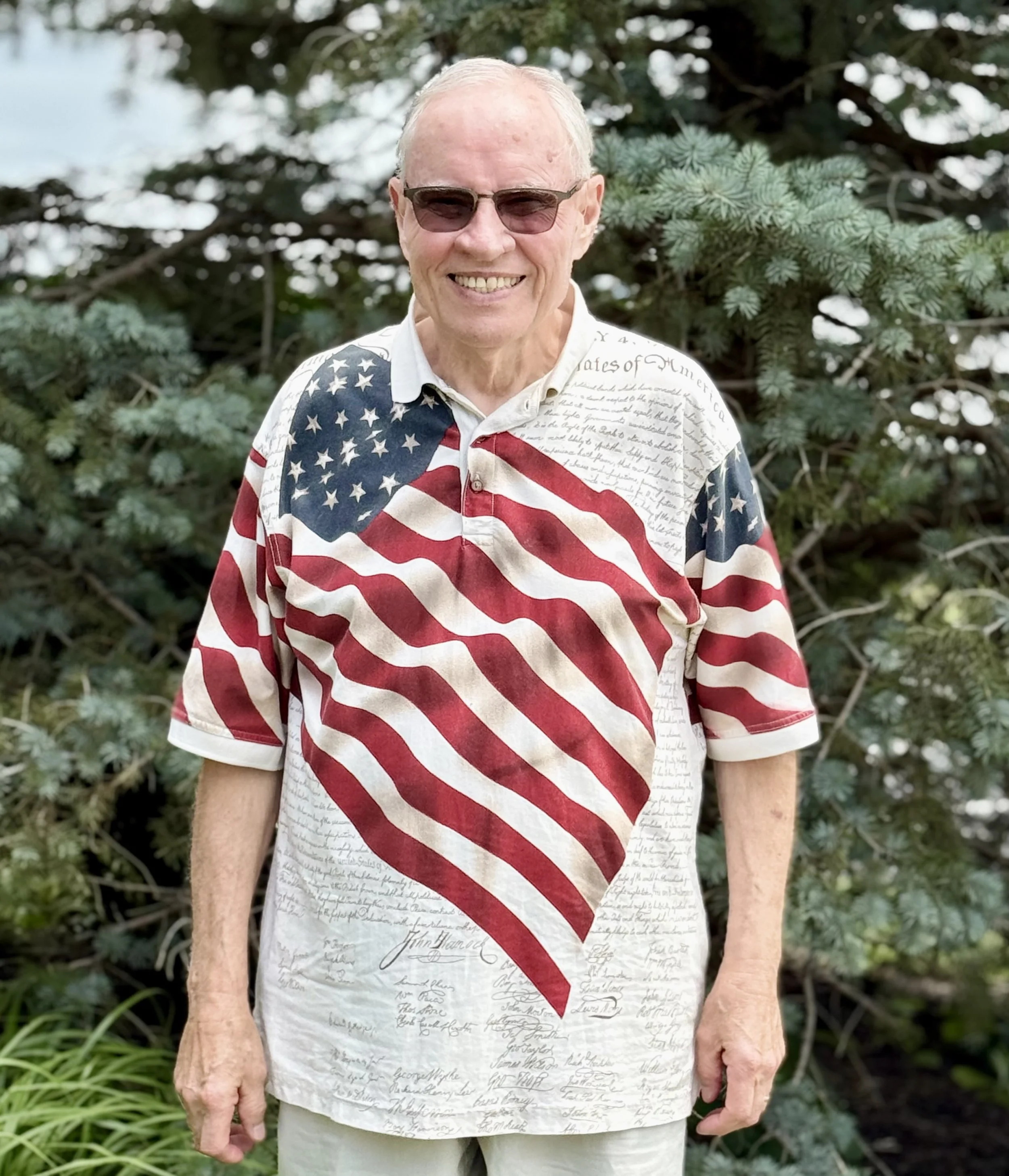 A smiling elderly man with gray hair and glasses, wearing a dark blazer and a red-striped shirt, against a dark background.