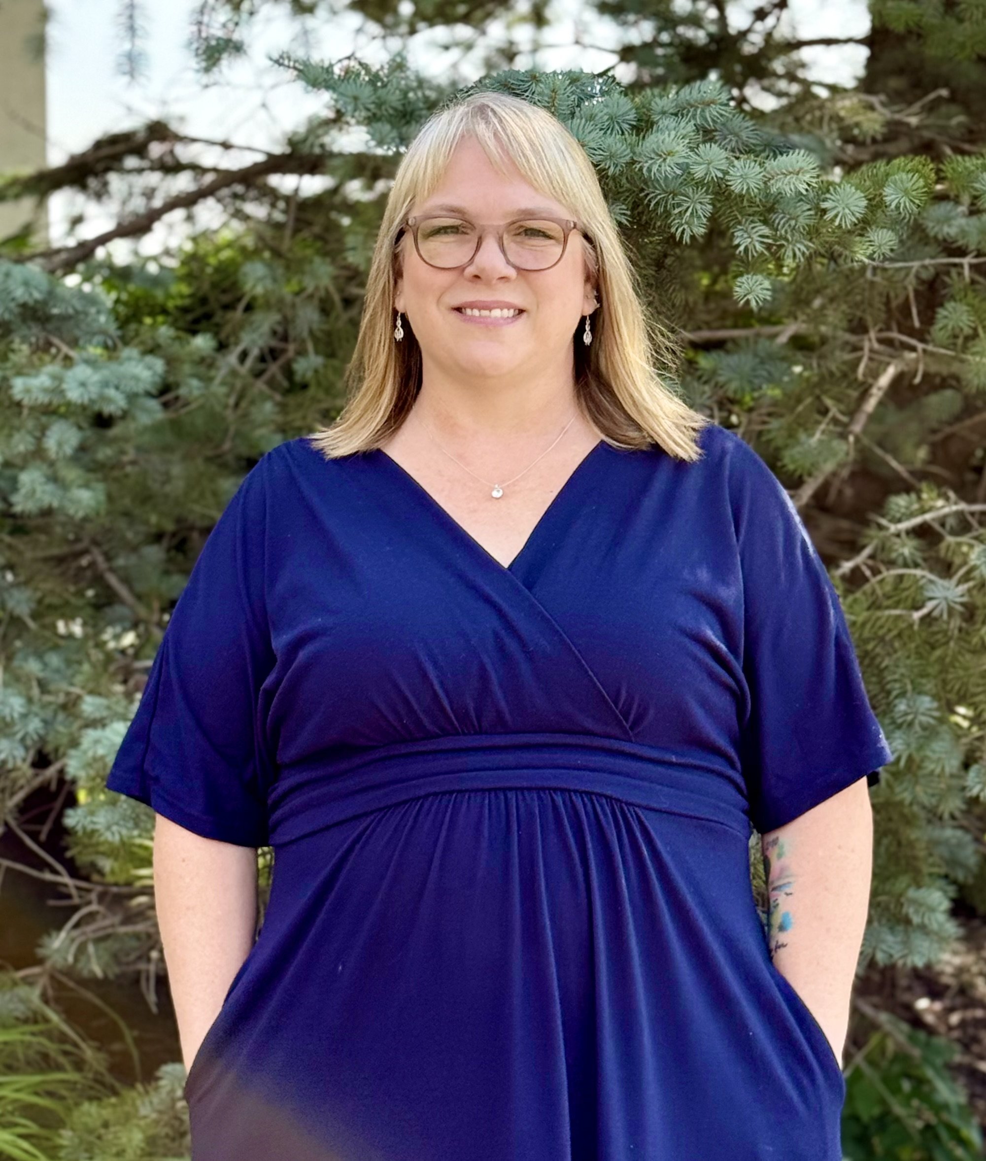 A woman with long blonde hair, glasses, and a floral dress smiling at the camera.