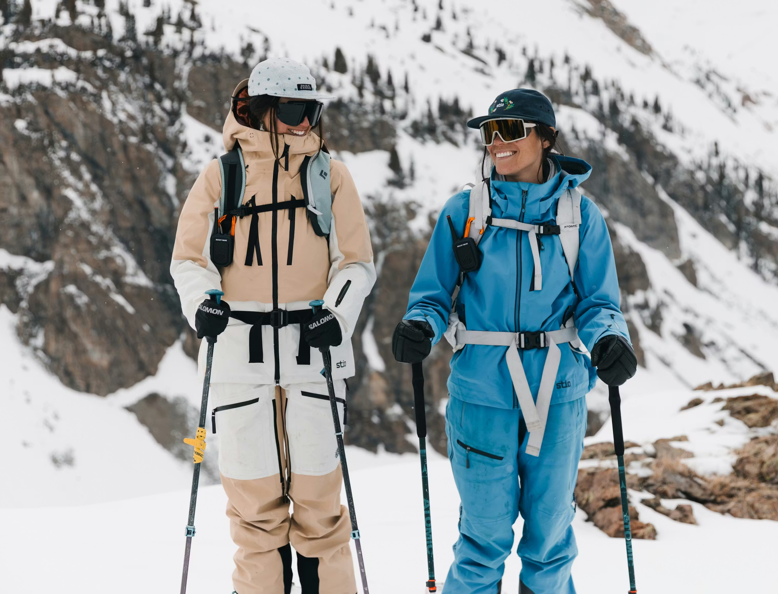 Two women in winter gear with snow-covered mountains in the background, smiling, holding ski poles, ready to ski or hike in a snowy landscape.