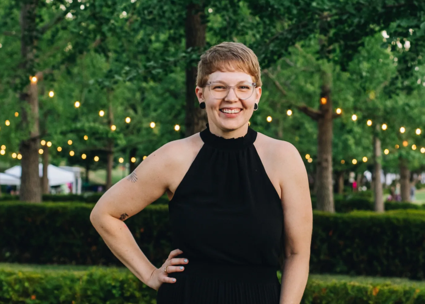 A smiling woman with short hair, glasses, and tattoos outdoors in a park with string lights and trees in the background.