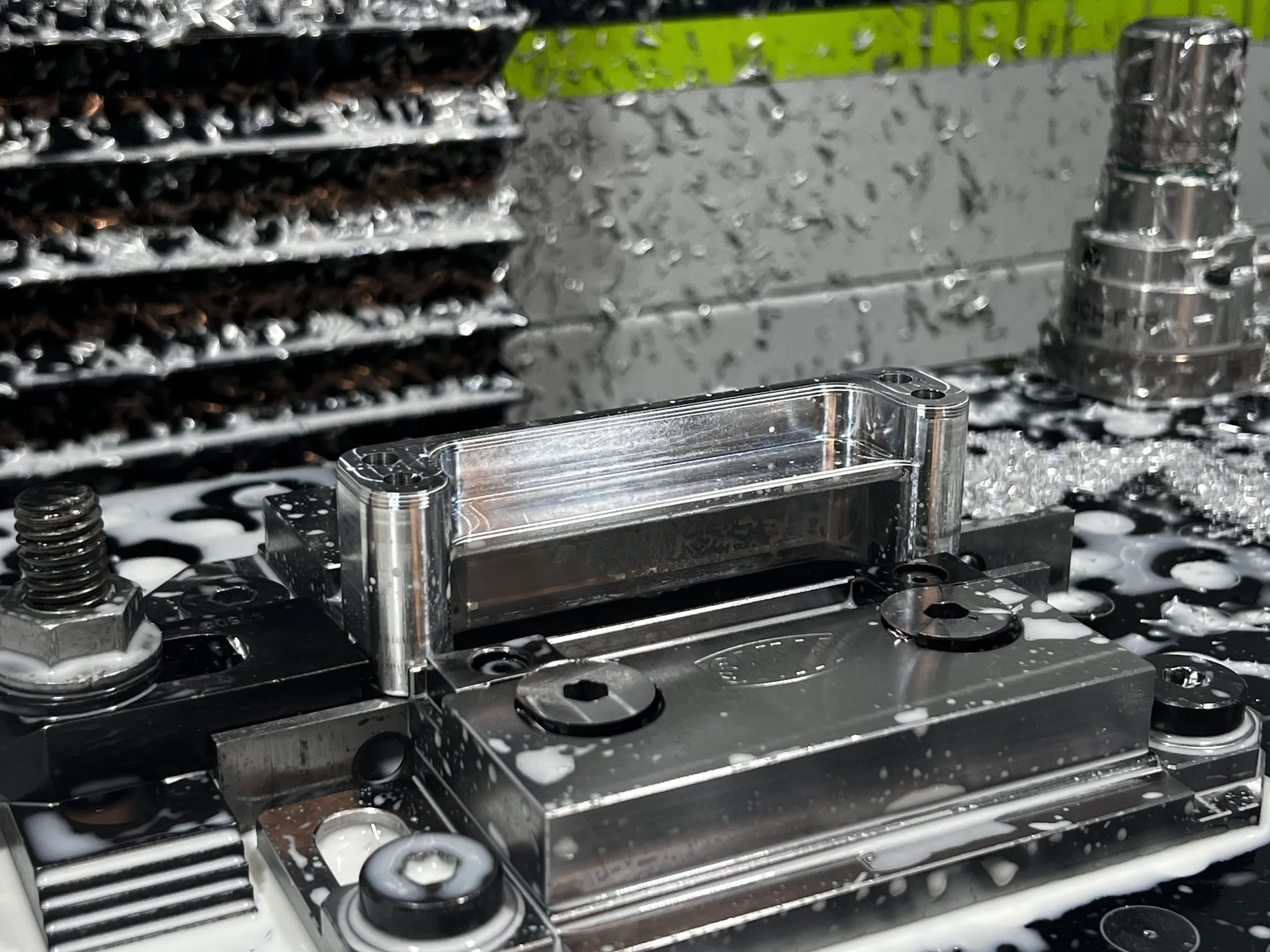 Close up photo of the 20:10 block being machined, the product is held in a vice on the cnc machining bed with coolant and metal chips scattered around.