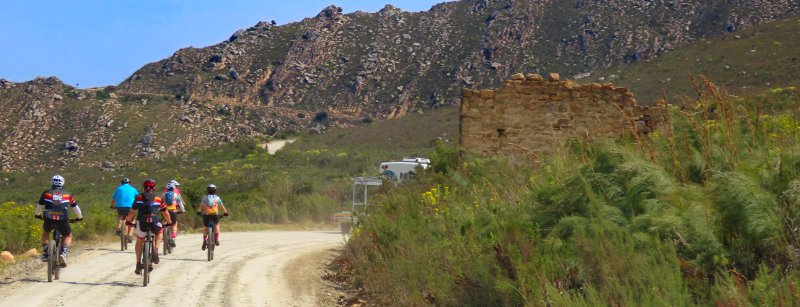 Six cyclists riding along a dirt road in a mountainous area with green vegetation and ruins on the side.