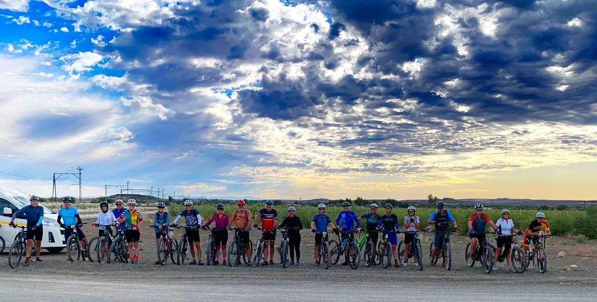 A group of 15 people with bicycles standing in a line outdoors on a dirt road, wearing helmets and sports attire, with a partly cloudy sky and some distant power lines in the background.