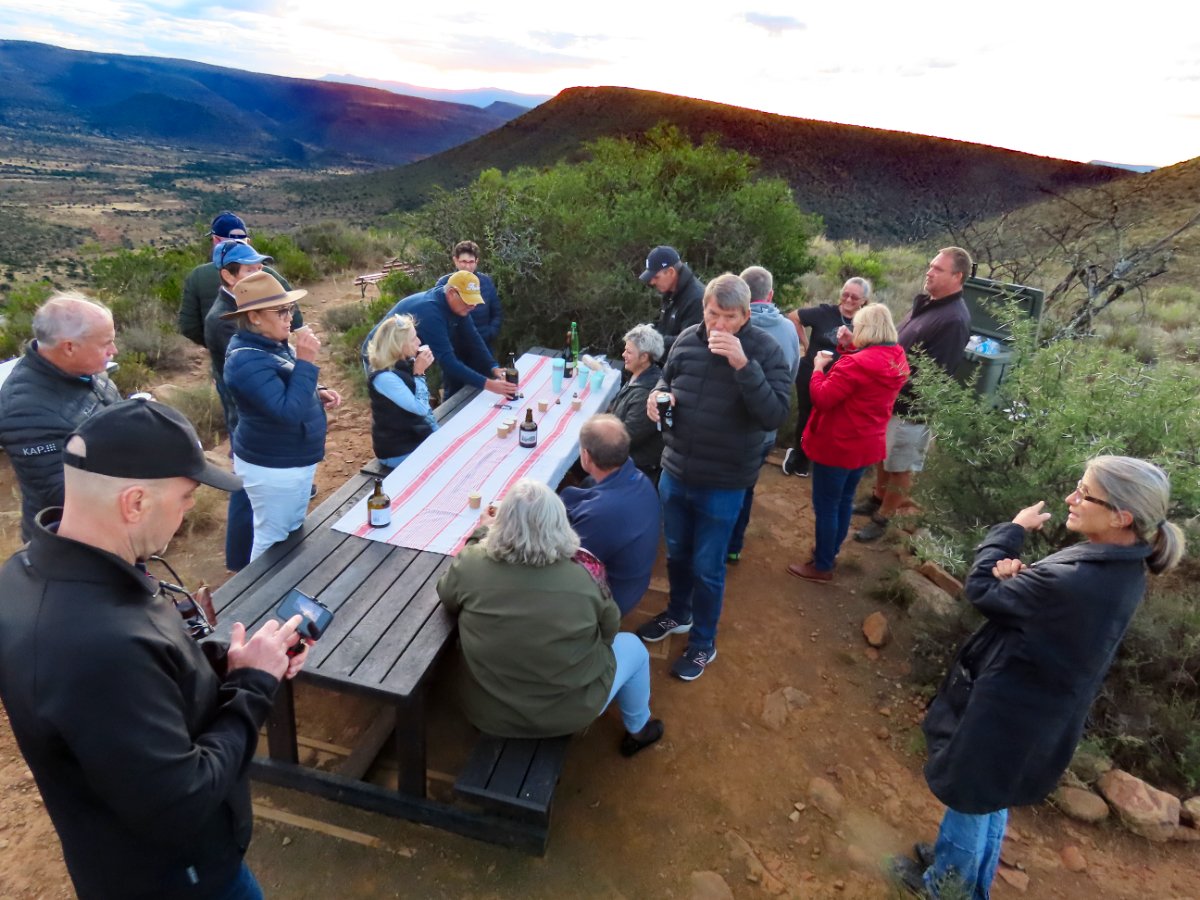 Group of people gathered outdoors on a dirt trail, some sitting at a picnic table with drinks and snacks, with desert landscape and mountains in the background during sunset.