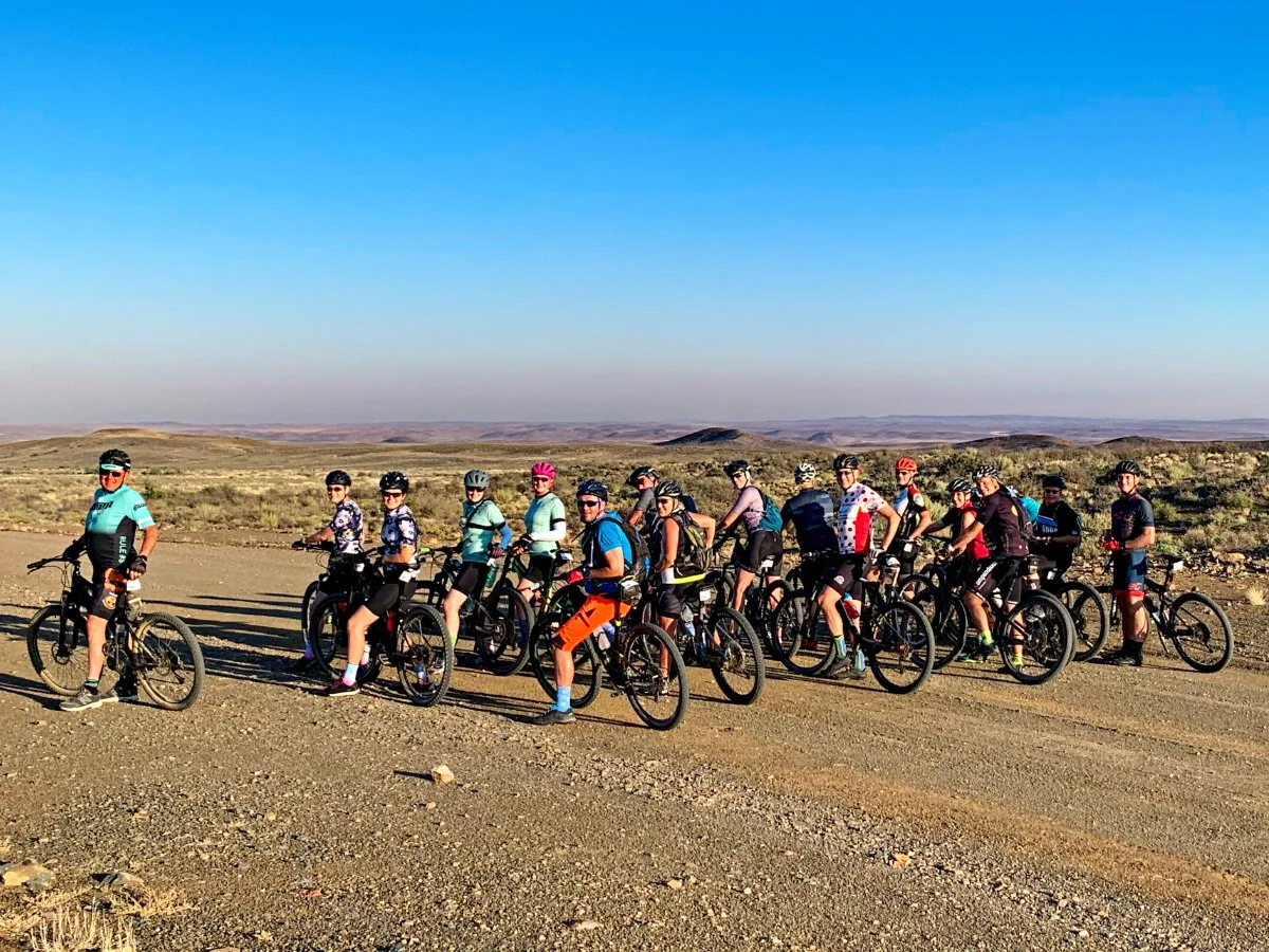 Group of cyclists riding on a dirt road in a desert landscape with hills in the distance under a clear blue sky.