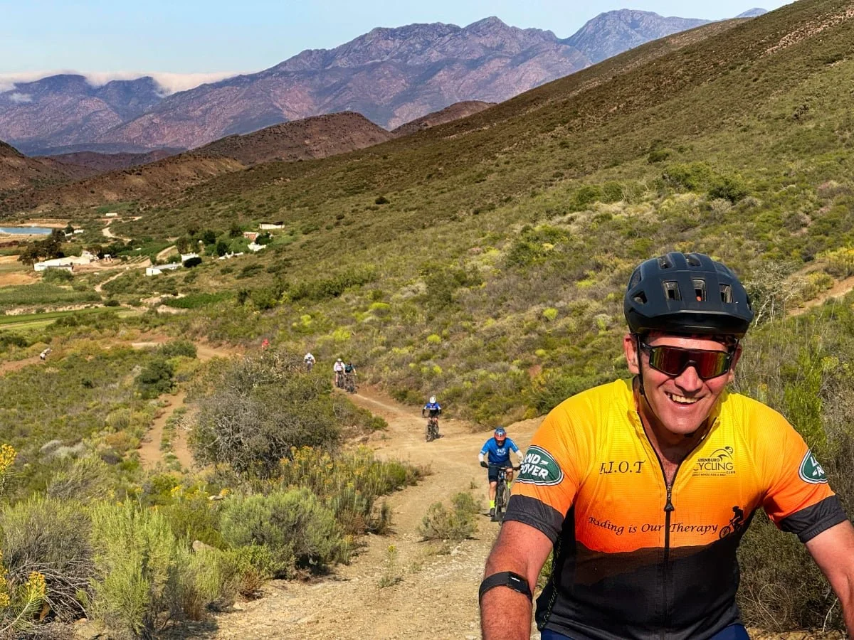 A group of mountain bikers riding on a dirt trail in a mountainous landscape with green shrubs and bushes, with mountains in the background, one man in the foreground wearing a black helmet, sunglasses, and a yellow-orange cycling jersey smiling.