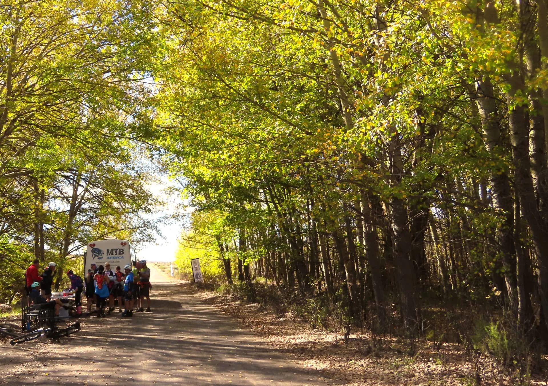 A group of cyclists gathered on a shaded dirt trail in a forest, with a van labeled 'MTB Africa' nearby and a person in a wheelchair on the ground, surrounded by trees with green and yellow leaves.
