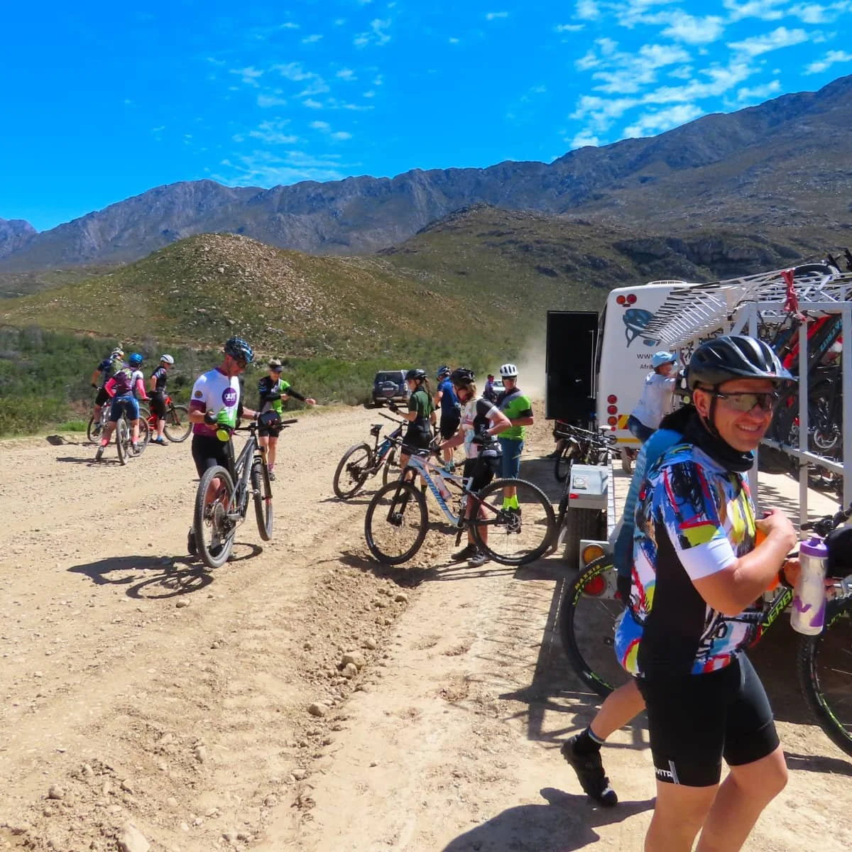 Group of mountain bikers at a trailhead with mountains in the background, some preparing their bikes and others socializing, under a clear blue sky.