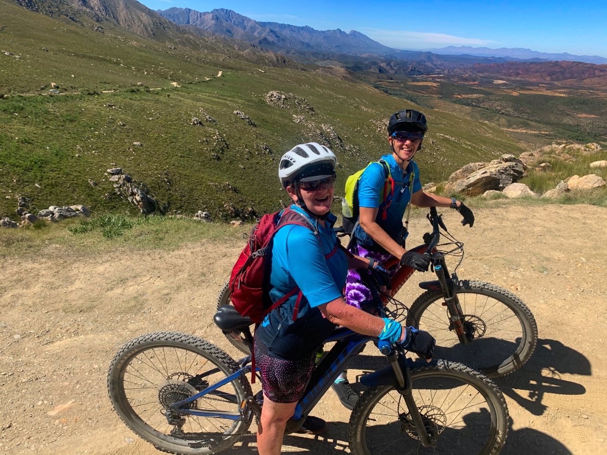 Two women in mountain biking gear smiling on dirt trail with mountains and open landscape in background.