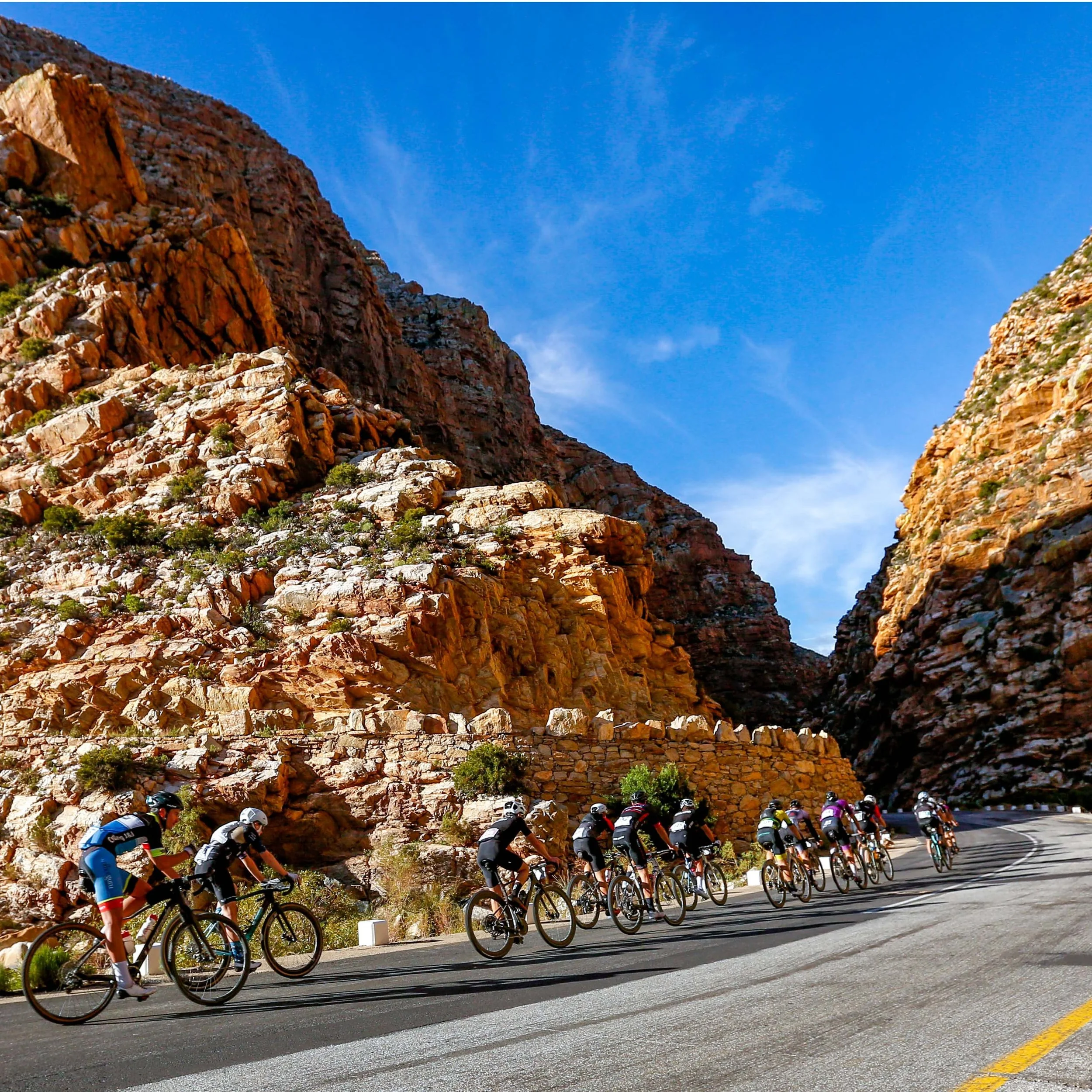 A group of cyclists riding on a mountain road with a rocky cliff on one side and a canyon on the other, under a blue sky.