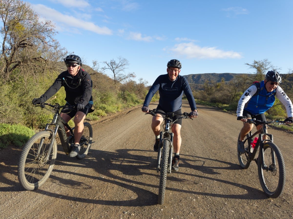 Three men riding mountain bikes on a dirt trail in a desert landscape with sparse trees and hills in the background.