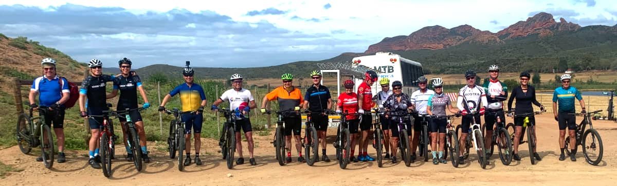 Group of cyclists with mountain bikes standing on a dirt path in a scenic outdoor area with mountains in the background.
