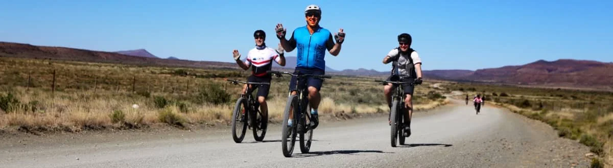 Three people riding mountain bikes on a dirt road in a desert landscape with hills in the background, one person in a blue shirt and helmet waving at the camera.