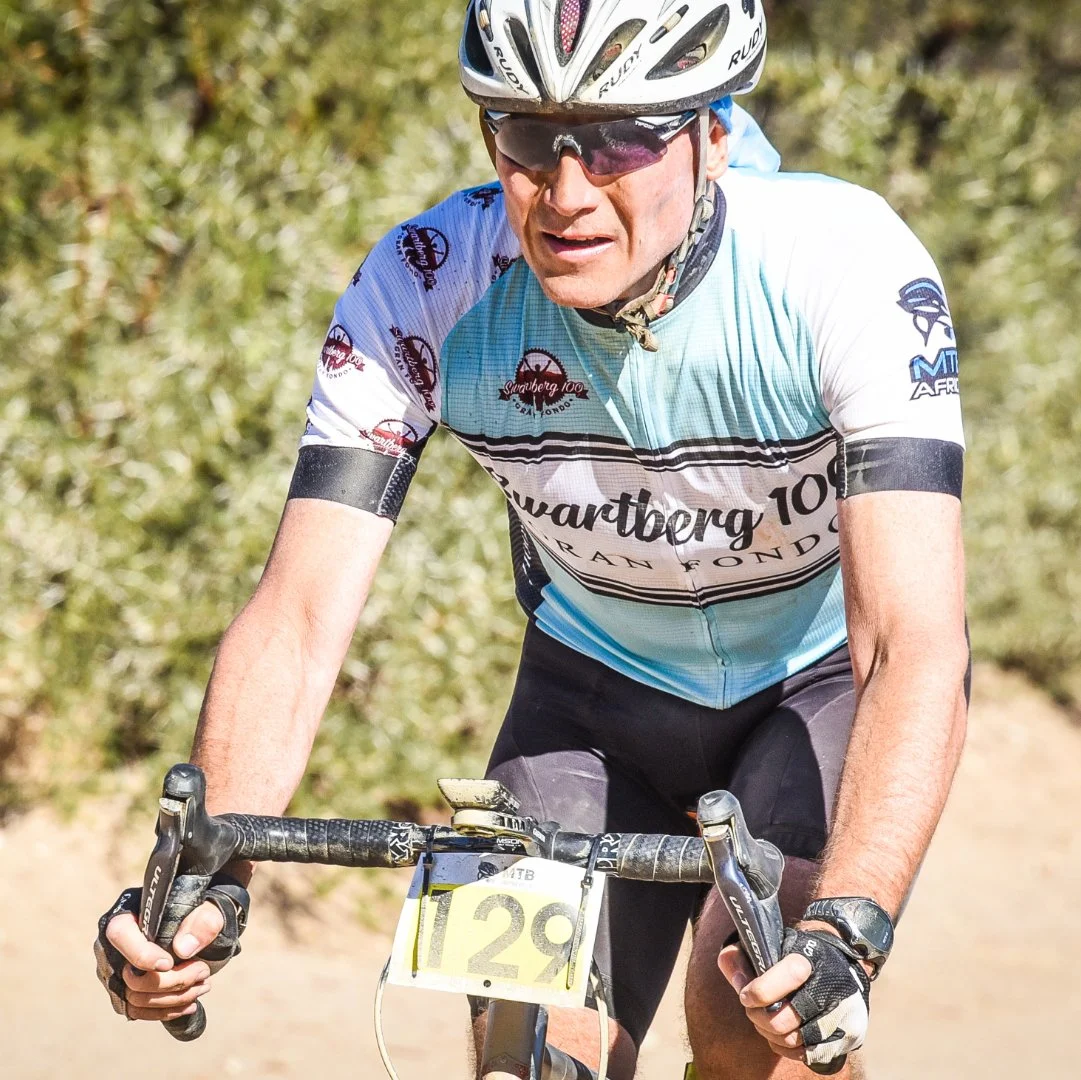 Male cyclist wearing a blue and white racing jersey, black shorts, a helmet, and sunglasses, riding on a trail with greenery in the background.