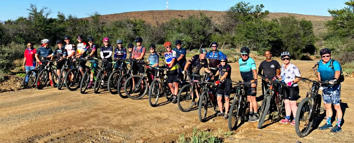 Group of people lined up with mountain bikes on a dirt trail in a rural area with sparse trees and hills in the background.
